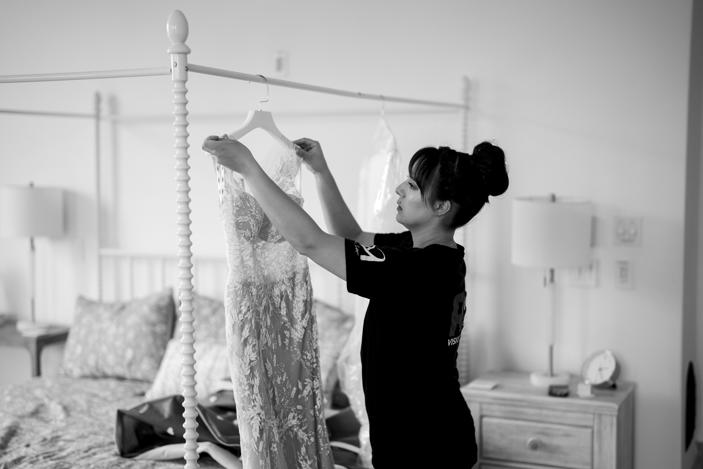 In a softly lit bedroom at Oxbow Estate, a woman holds up a lace wedding dress on a hanger. The four-poster bed, flanked by two-night stands and covered with a patterned duvet, adds to the timeless elegance captured in this black-and-white wedding photography scene.