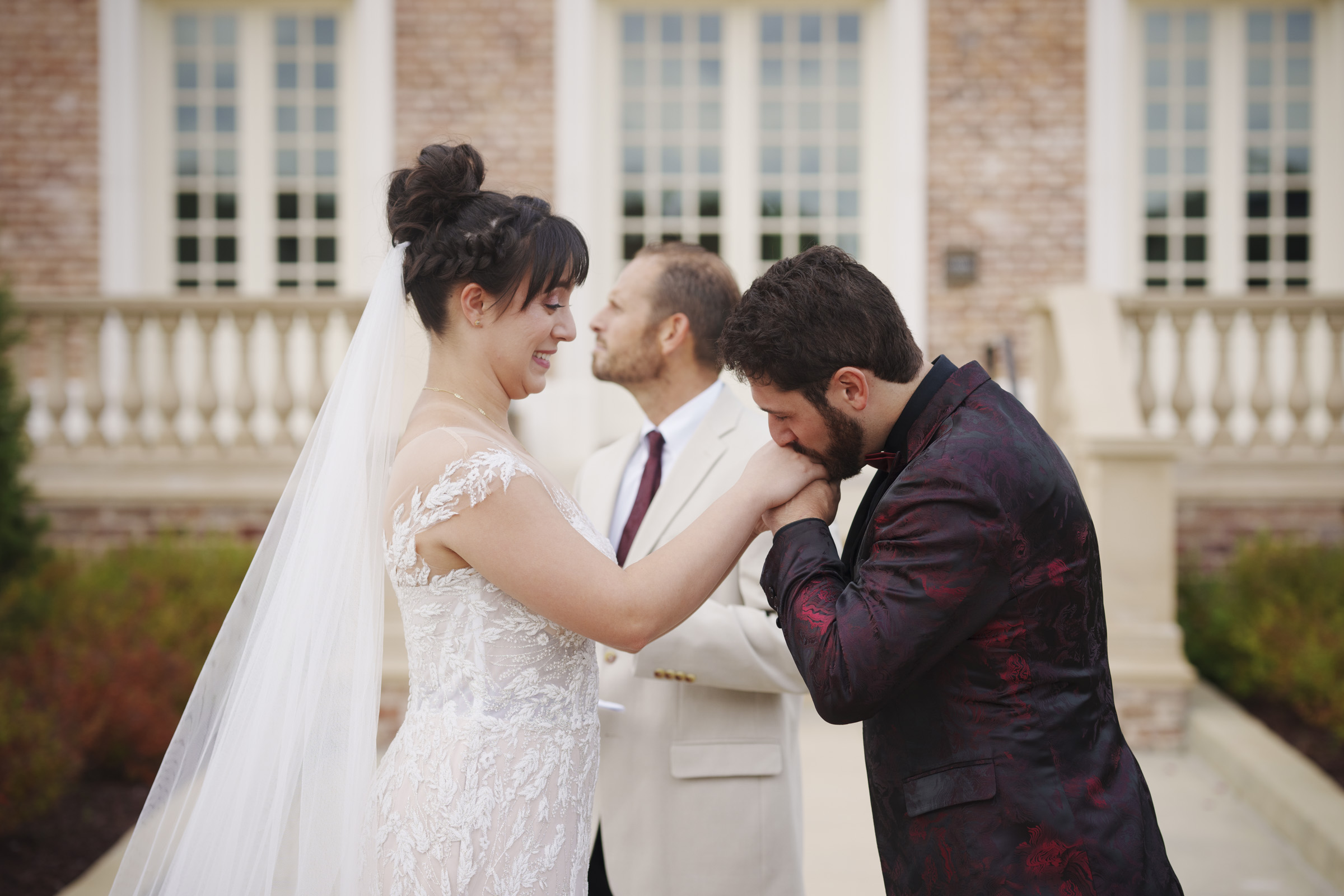 A bride in a white lace dress and veil smiles as the groom, wearing a dark suit, kisses her hand. Captured by Oxbow Estate Wedding Photography, they stand outside in front of a brick building with tall white windows. A man in a light suit stands behind them, looking upward.