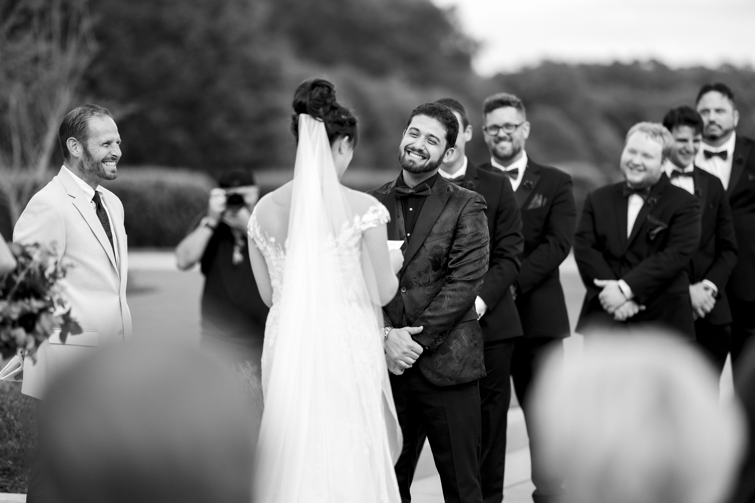 A black and white photo captures an Oxbow Estate wedding ceremony. The groom, smiling, faces the bride in a white gown and veil. They stand outdoors, with groomsmen and a photographer subtly framing this enchanting moment.