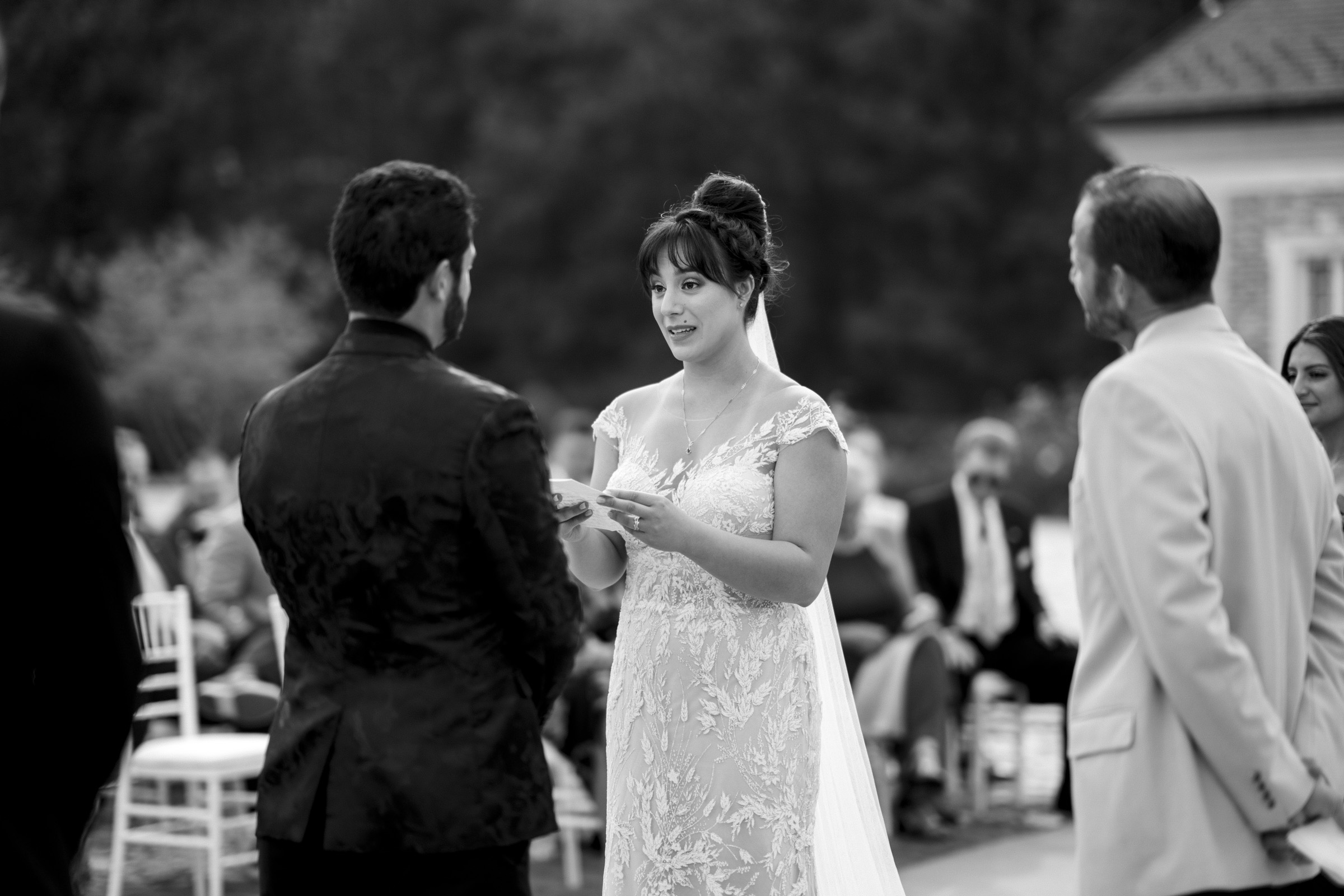 A bride in a lace gown reads vows to her groom, who is wearing a dark suit, captured beautifully by Oxbow Estate Wedding Photography. They stand outdoors with guests seated in the background, while one person observes. The enchanting scene is elegantly portrayed in black and white.