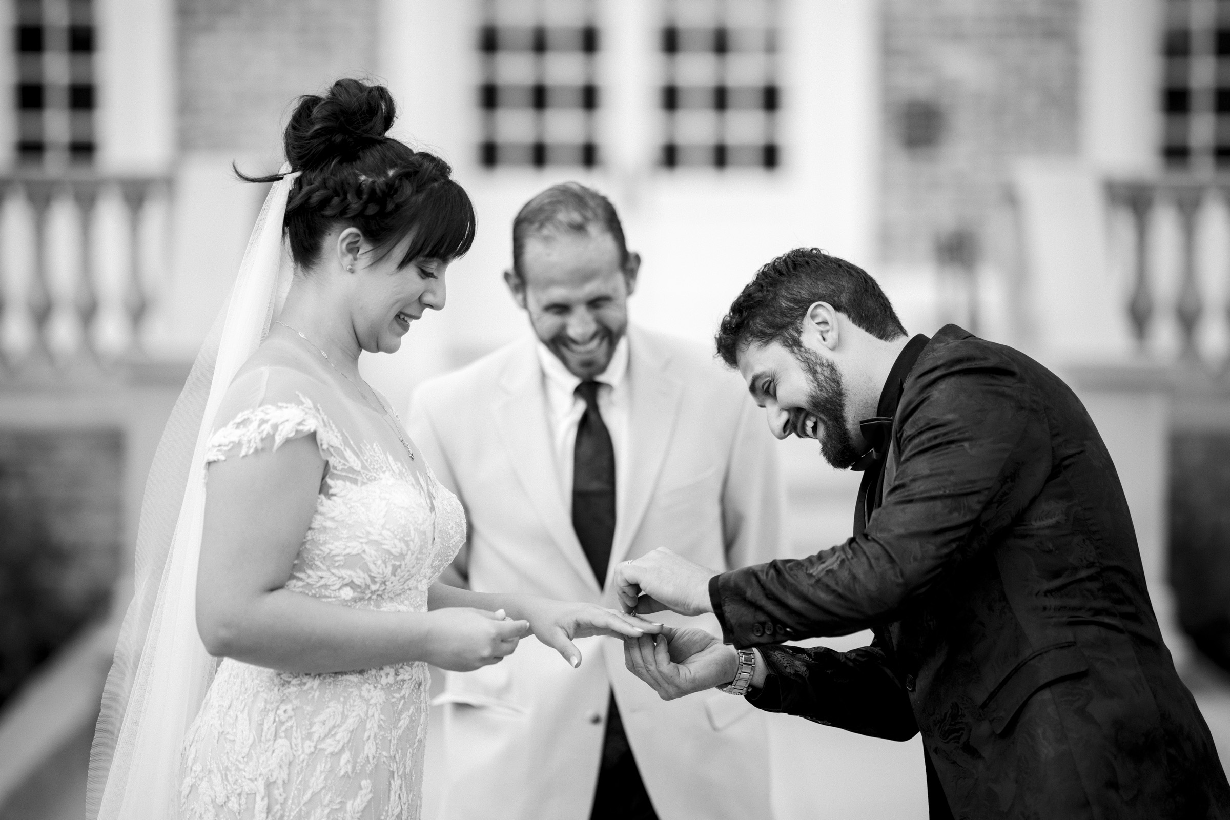 At Oxbow Estate, a bride and groom exchange rings during a wedding ceremony. The bride, in a lace gown, smiles as the groom, in a dark suit, slips a ring on her finger. The officiant, in a light suit, stands behind them, smiling. Captured beautifully in black and white photography.