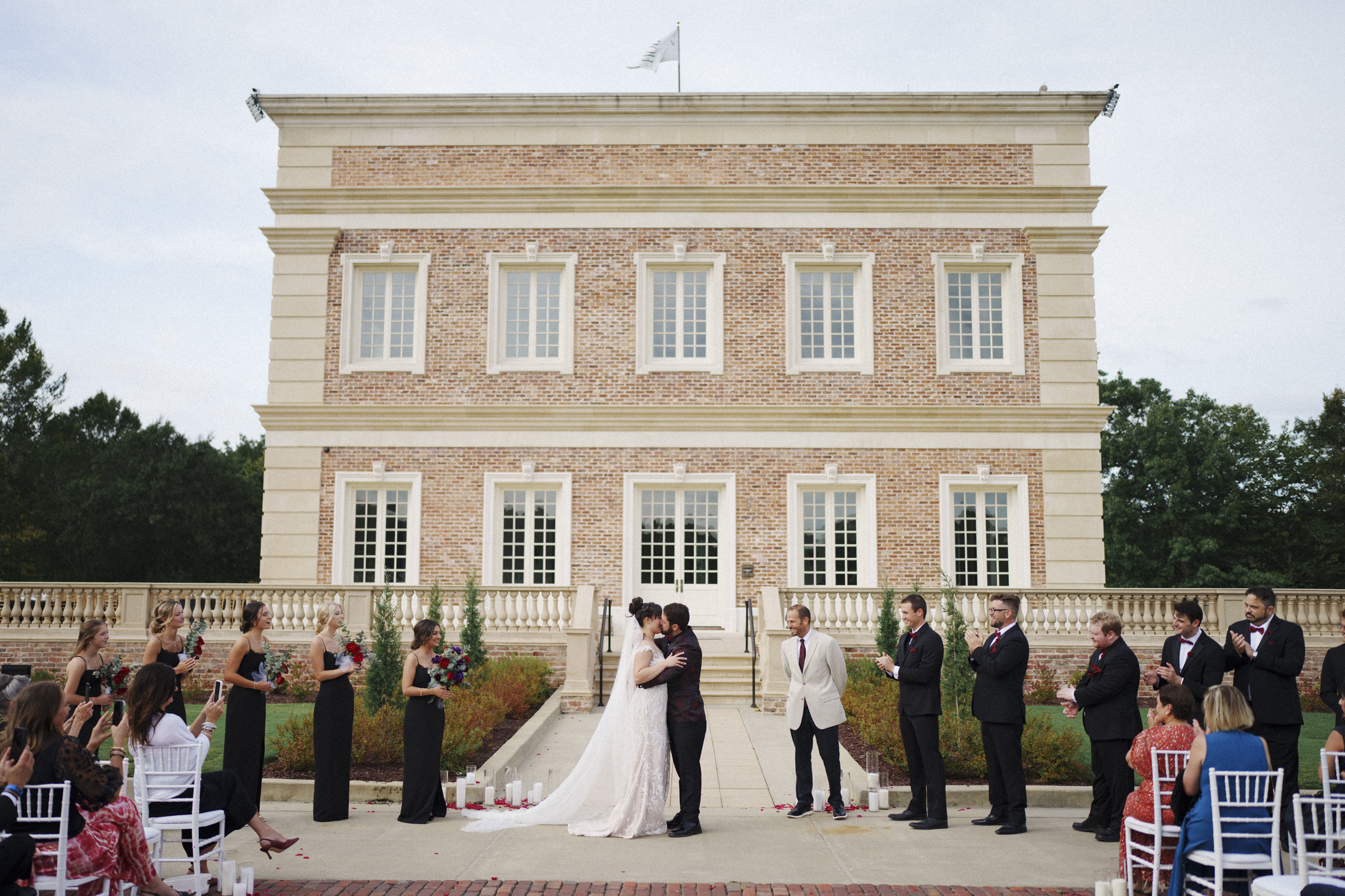 At Oxbow Estate, a bride and groom share a kiss in front of the grand, ornate building during an outdoor wedding ceremony. The wedding party in formal attire stands on either side, with guests seated facing the couple amid lush trees and greenery—a perfect scene for wedding photography.