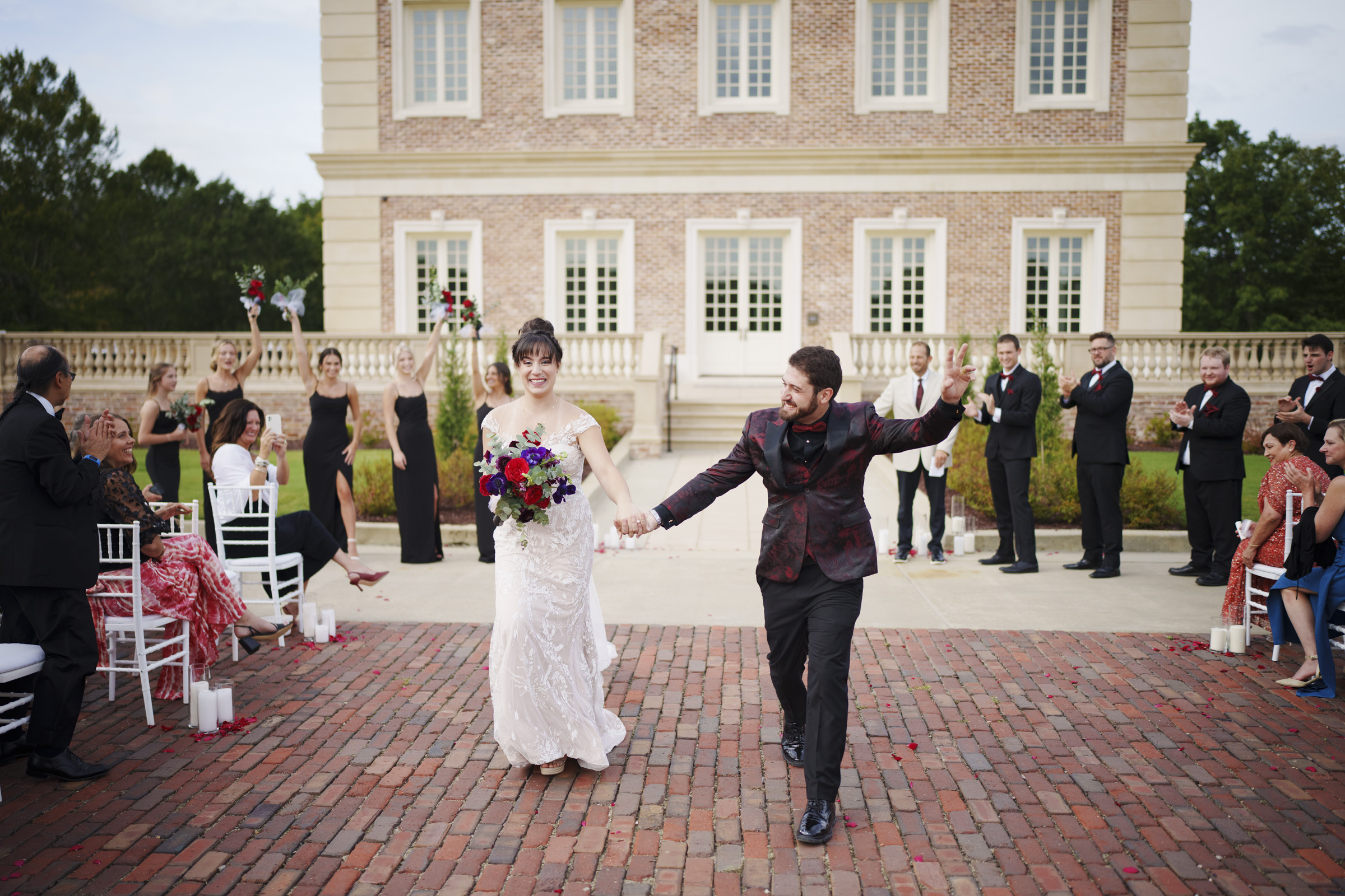 A bride and groom joyfully walk down a brick pathway at the picturesque Oxbow Estate. The bride holds a bouquet of red and purple flowers, capturing the essence of the moment. Guests, dressed in formal attire, stand and cheer on either side, making this wedding photography truly memorable.