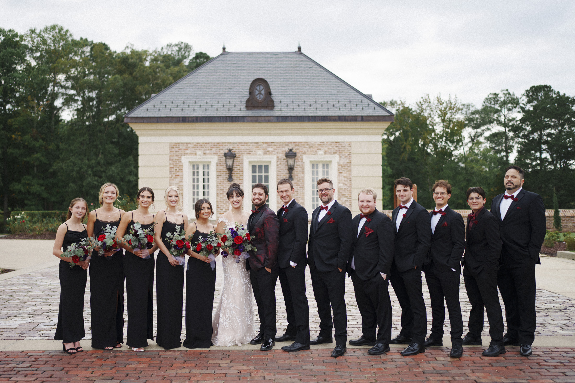 A wedding party stands in front of a small building at Oxbow Estate. The group features bridesmaids in black dresses and groomsmen in black suits with red accents. The bride shines in a white gown, while the groom sports a suit with a bow tie. Lush trees provide a beautiful backdrop, perfect for wedding photography.