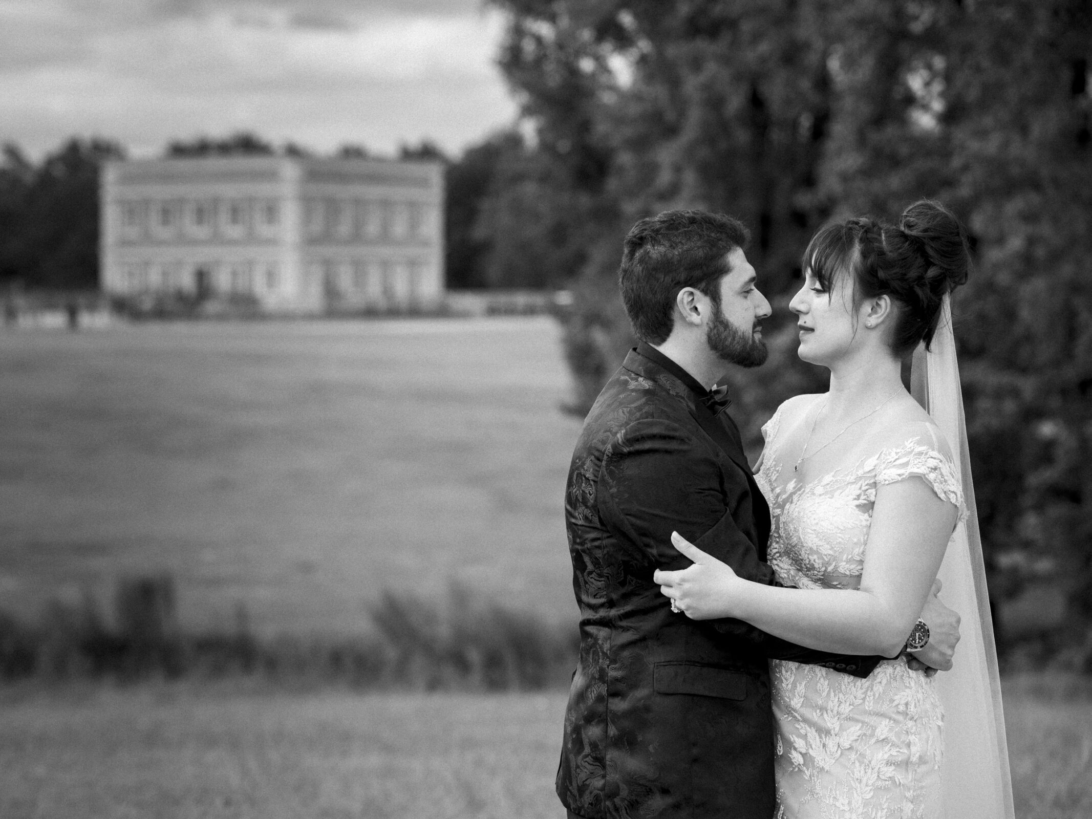 A black and white photo captures a couple in wedding attire embracing outdoors at the picturesque Oxbow Estate. They stand on grass with a large building and trees in the background, looking warmly at each other, perfectly encapsulating the essence of estate wedding photography.