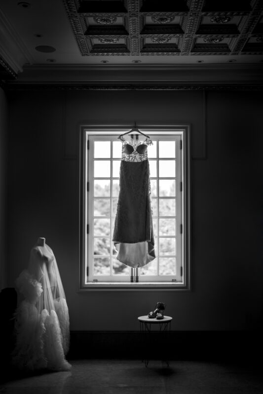 A monochrome image from Oxbow Estate Wedding Photography captures a wedding dress hanging in front of a large window. Beside it, on a mannequin, is another gown with voluminous fabric. Below, a pair of shoes rests on a small round table, while the ornate ceiling adds elegance above.