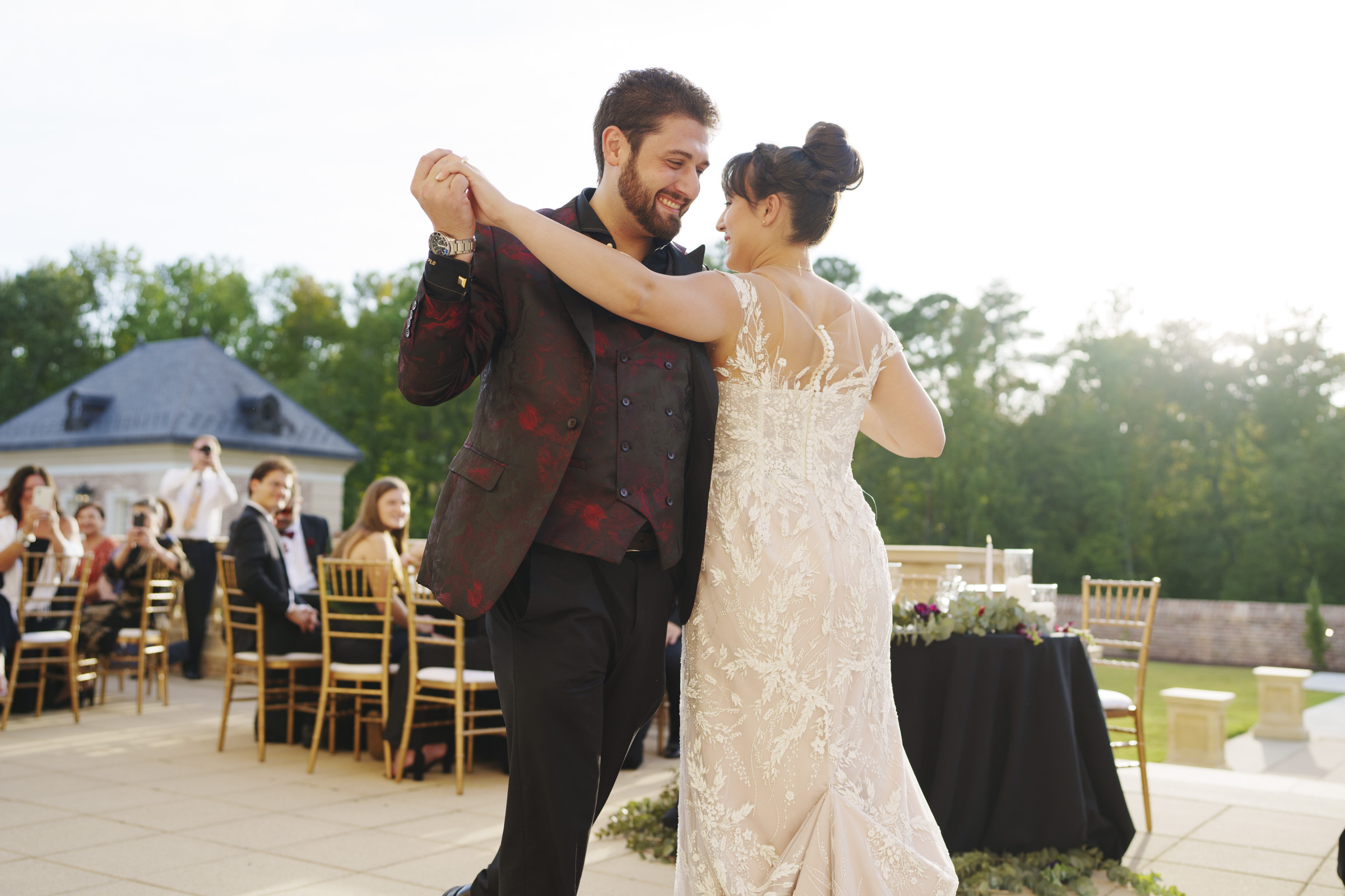 At an Oxbow Estate wedding, a couple dances gracefully outdoors. The groom dons a dark patterned suit, and the bride glows in a white lace dress. Guests seated nearby watch with smiles under a bright sky framed by trees, capturing picture-perfect moments of joy and romance.