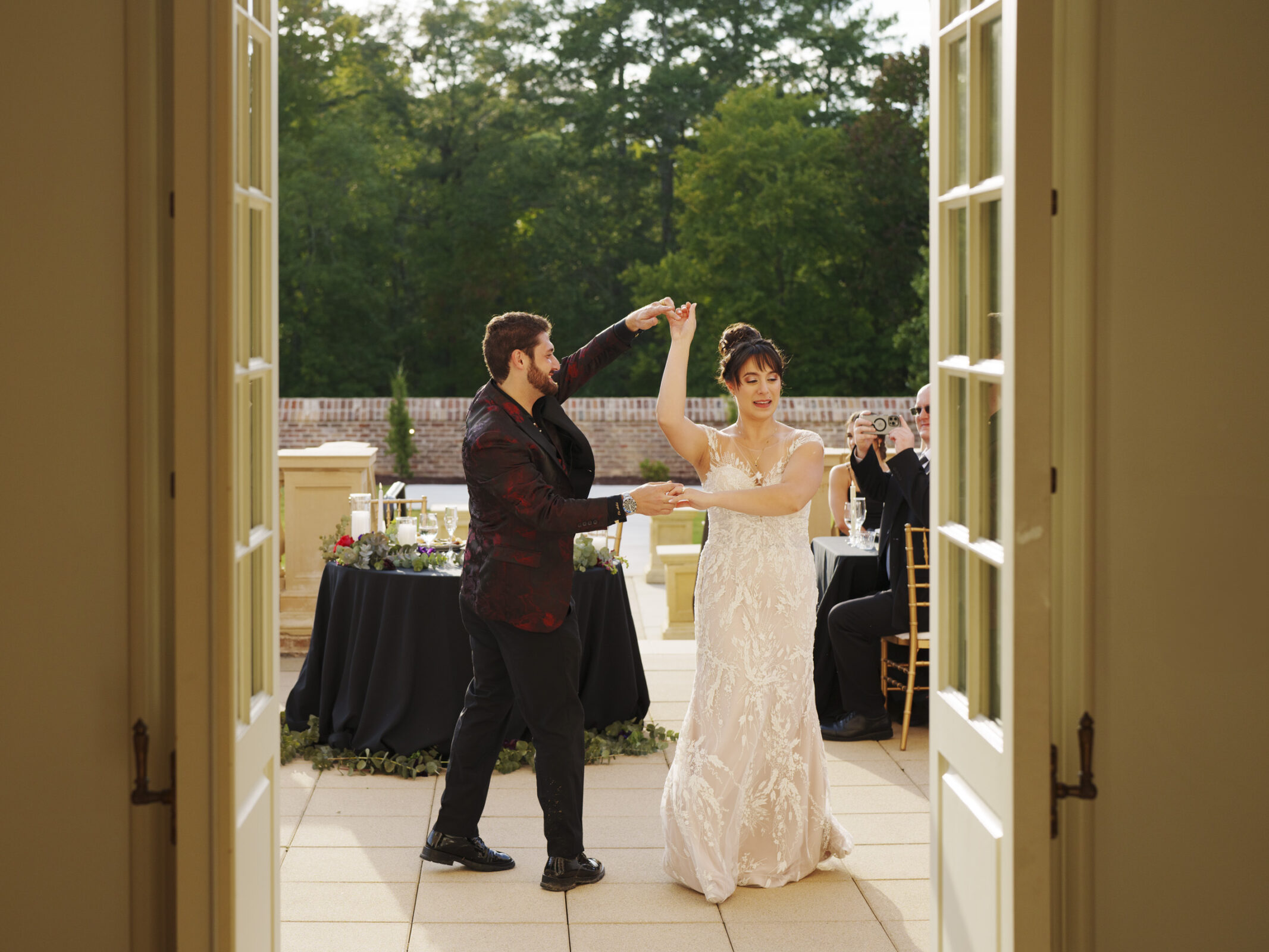 A bride in a white gown and a groom in a dark suit are dancing on an outdoor terrace at Oxbow Estate, surrounded by greenery. An open door frames the scene as a photographer captures the moment beside a round table adorned with a floral arrangement.