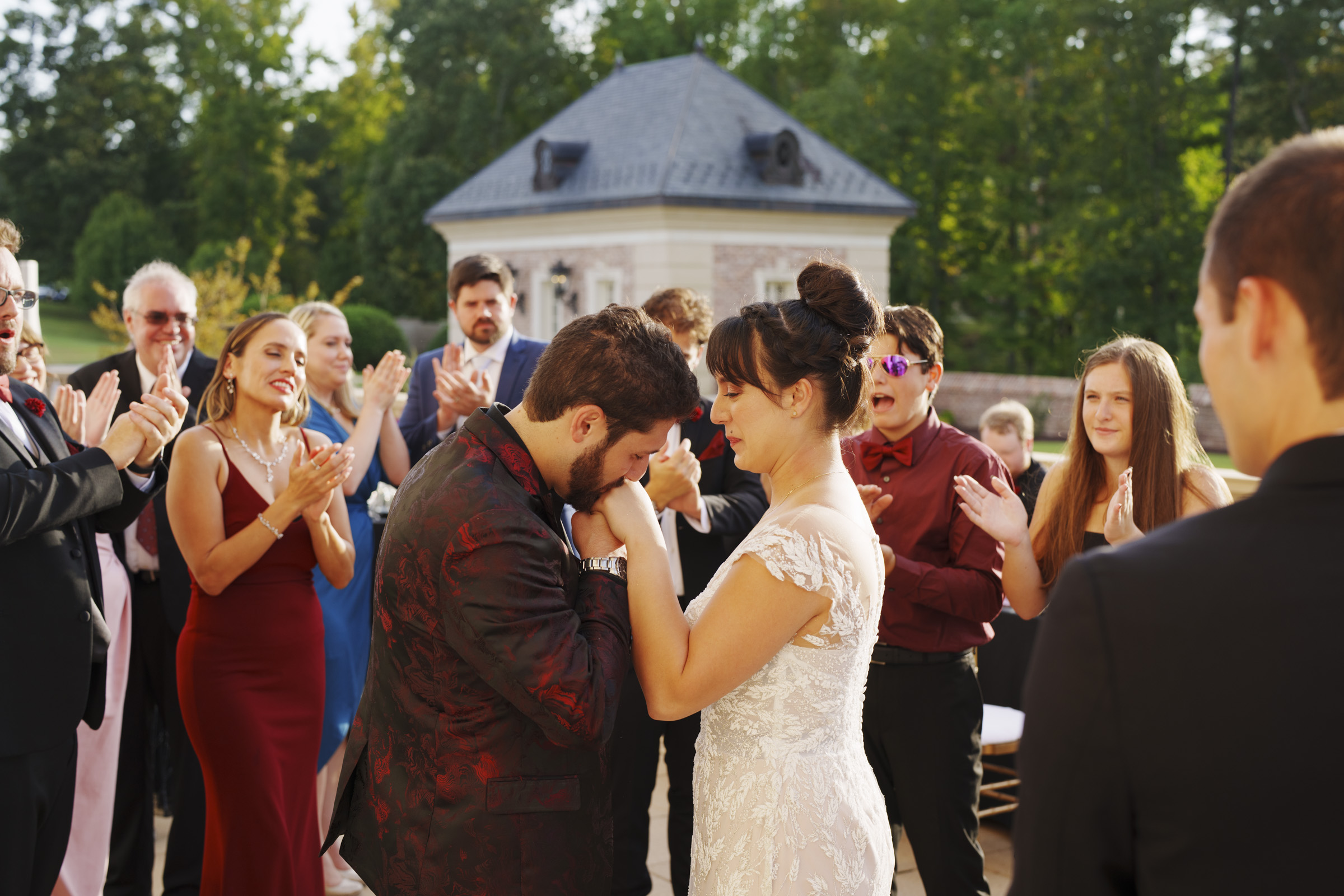 At the picturesque Oxbow Estate, a groom in a patterned suit tenderly kisses the bride’s hand during an outdoor wedding ceremony. The bride dazzles in her lace gown as guests in formal attire clap and smile. A charming building and lush trees provide the perfect backdrop for this unforgettable moment.