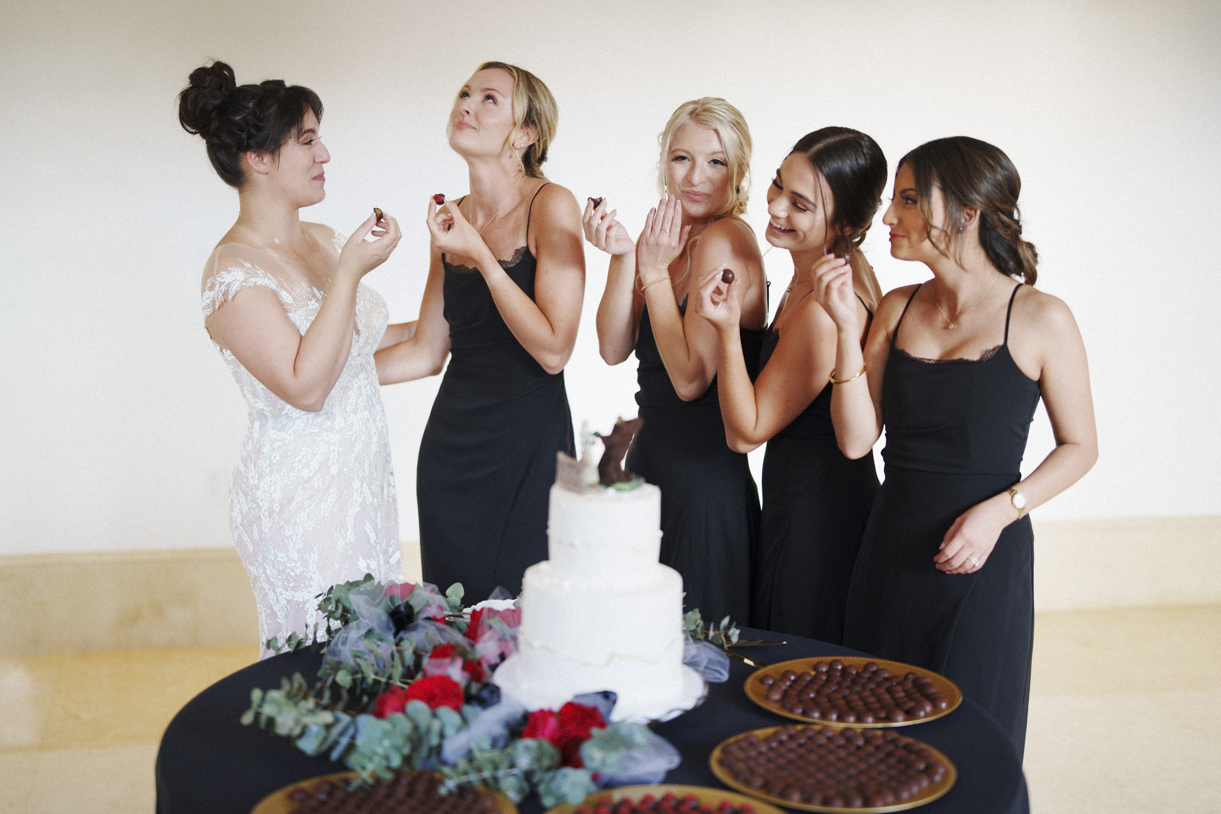 At an Oxbow Estate Wedding, a bride in a white dress and five bridesmaids in black dresses pose joyfully near a table adorned with a white cake and chocolates. With smiles as radiant as the decorative flowers, they playfully hold their sweet treats, captured perfectly in every photograph.