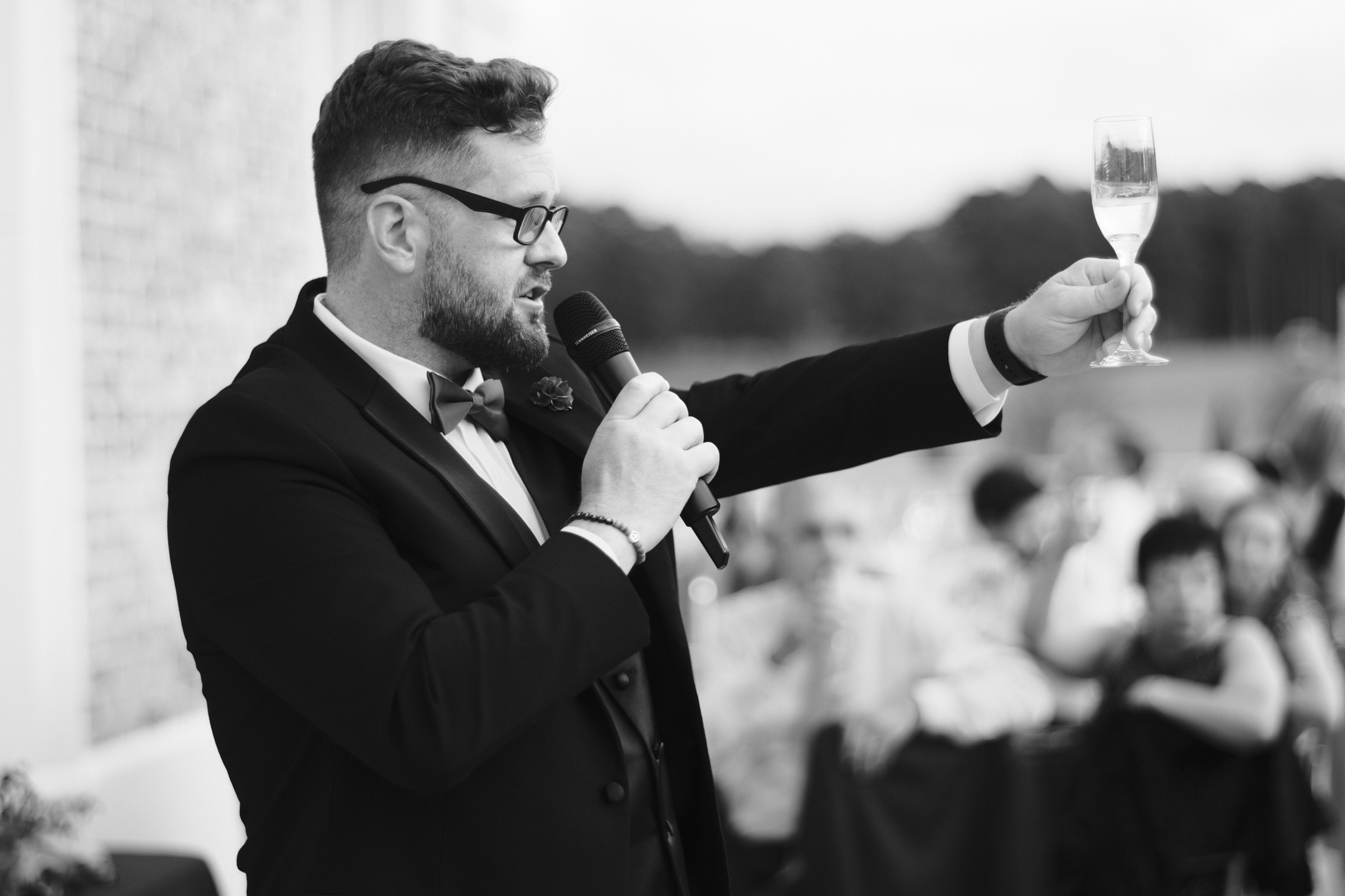 A bearded man in a suit holds a microphone and raises a glass in a toast during an outdoor event at Oxbow Estate. He stands before a seated crowd, with the sweeping field as his backdrop. This black-and-white moment perfectly captures the essence of wedding photography.