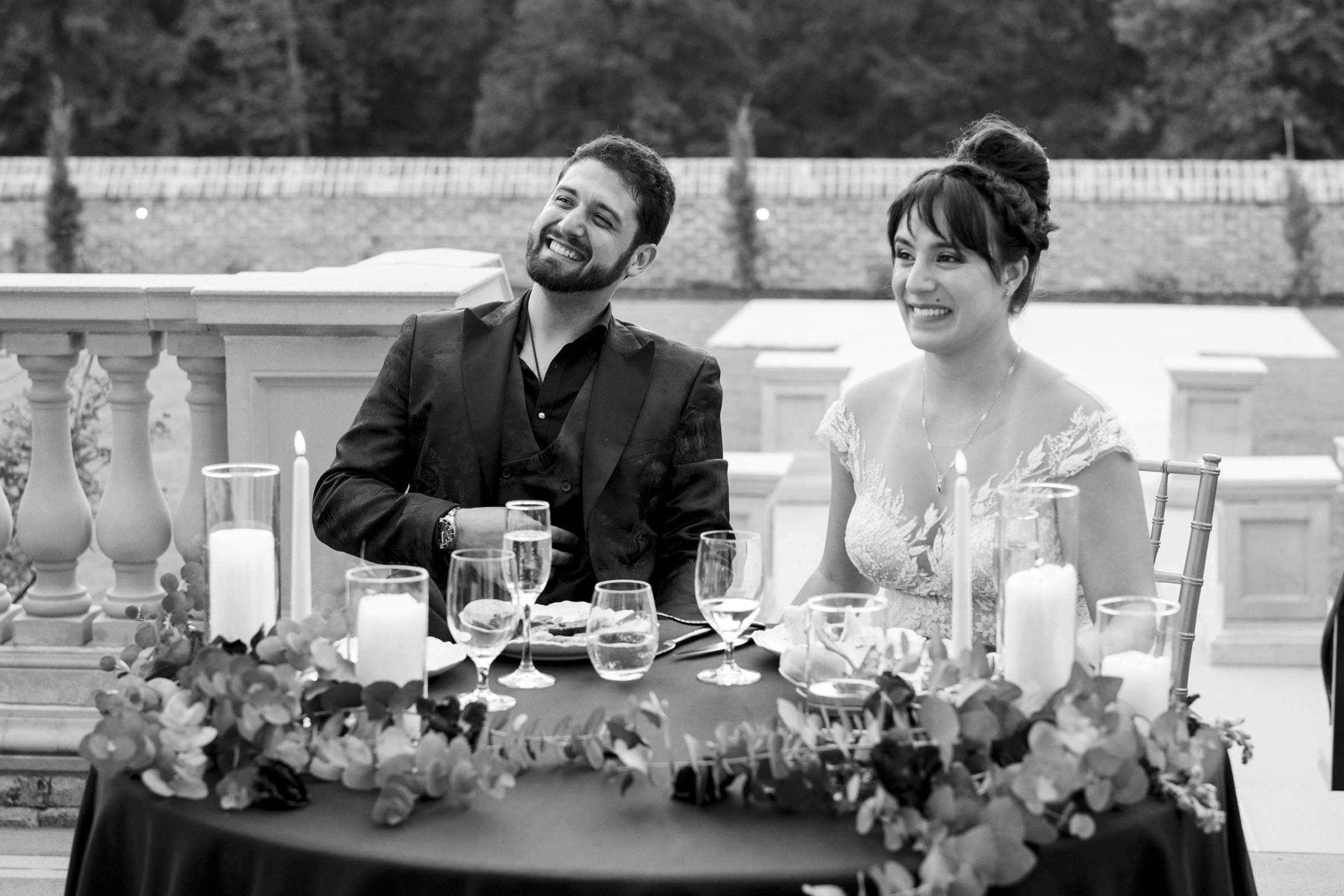 A smiling couple sits at a table adorned with flowers and candles at an elegant outdoor event. Captured by Oxbow Estate Wedding Photography, the scene features the man in a suit and the woman in a dress, with a stone railing and trees gracing the background.