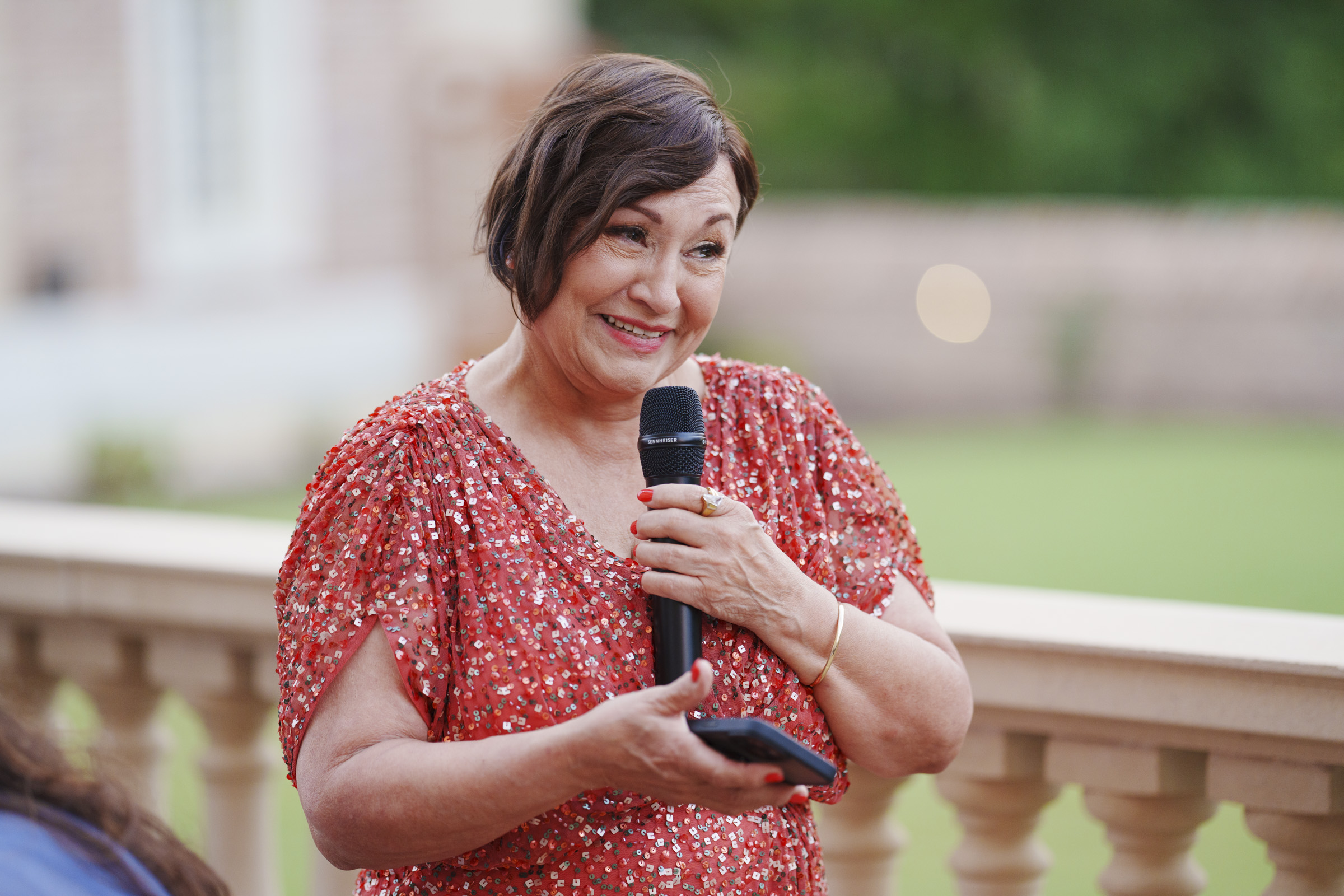 A woman in a red, sparkly dress smiles while holding a microphone and a phone, capturing memories at an Oxbow Estate Wedding. She stands outdoors near a balcony with a blurred green background, perfectly blending elegance and joy.