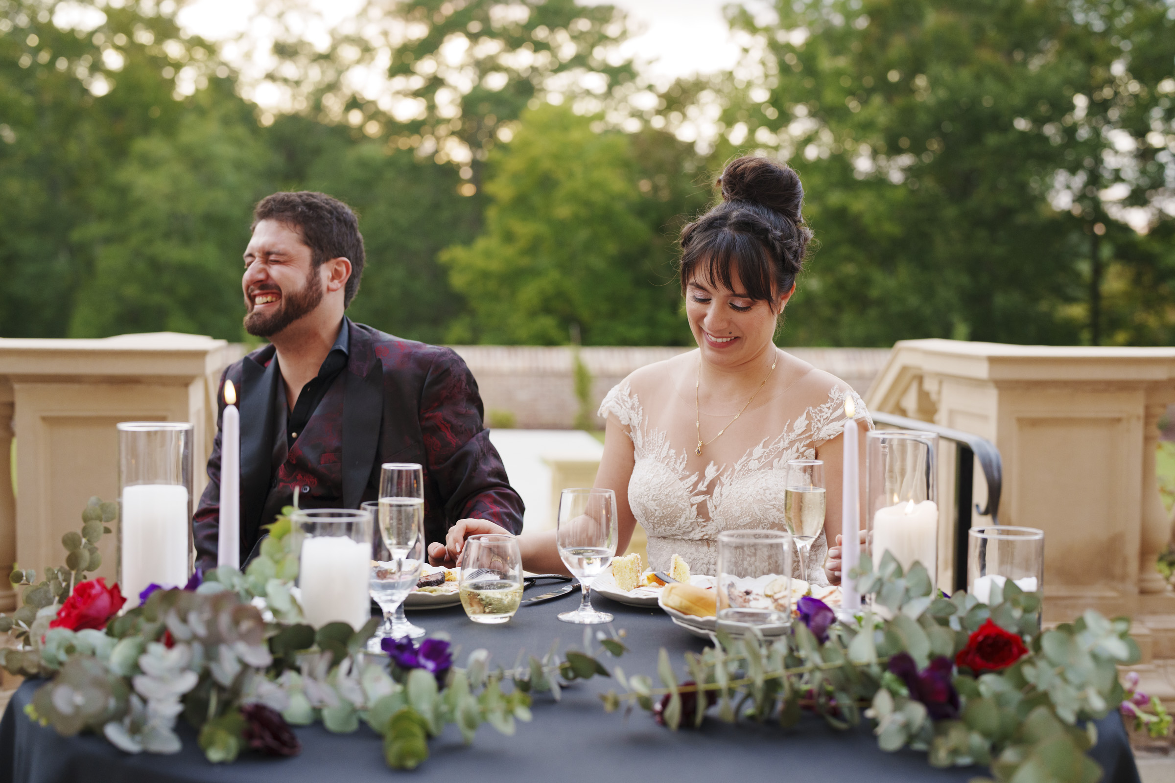 At an Oxbow Estate wedding, a bride and groom sit at an outdoor table adorned with flowers and candles. The bride smiles gently, looking down, while the groom bursts into laughter. Behind them, lush greenery and soft evening light create a magical backdrop for this special moment.