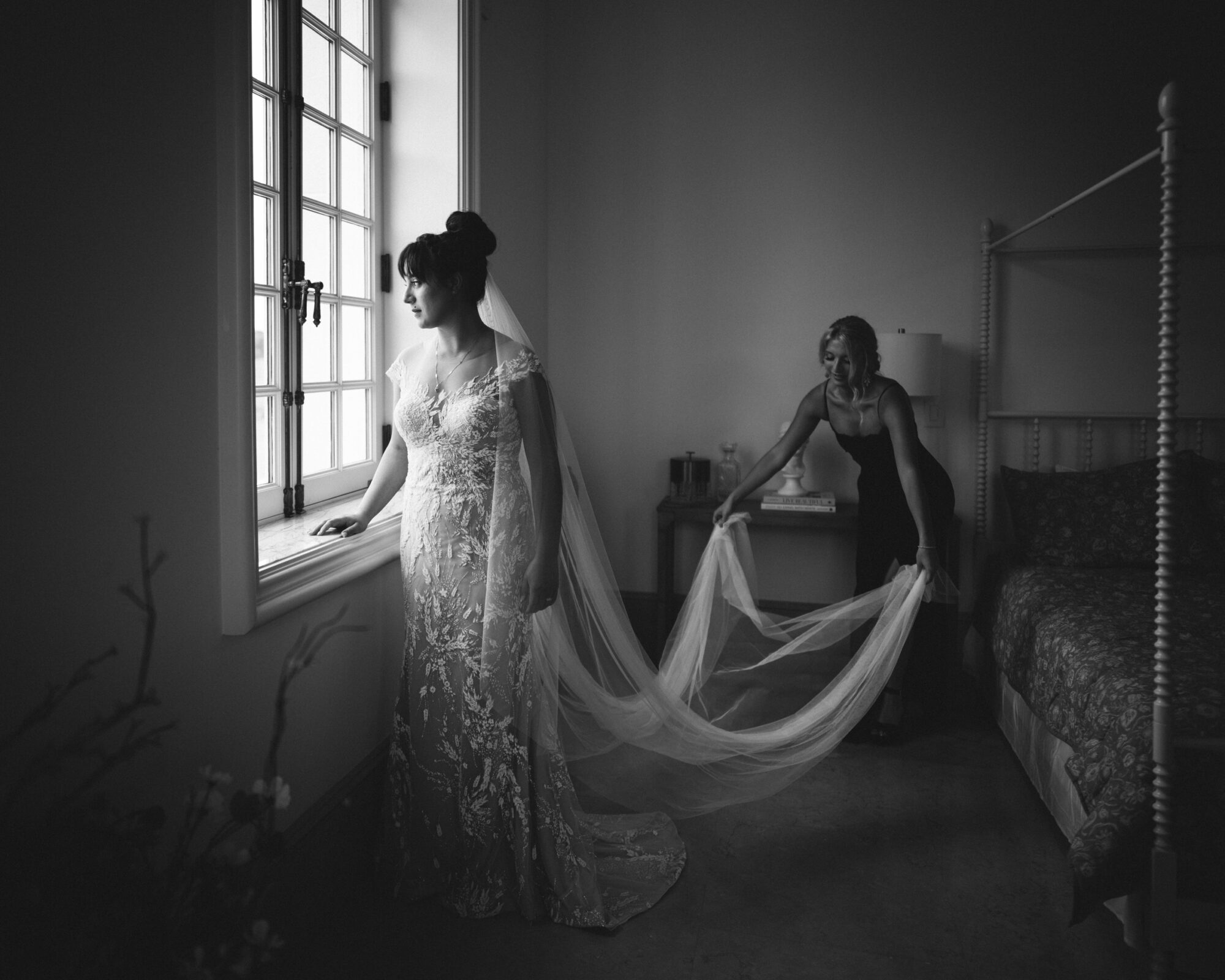 A bride in an ornate lace gown gazes out of a sunlit window at Oxbow Estate. Her veil flows gracefully, assisted by a woman in a dark dress. The room is softly lit, highlighting elegant details and creating a serene, intimate atmosphere, perfect for wedding photography.