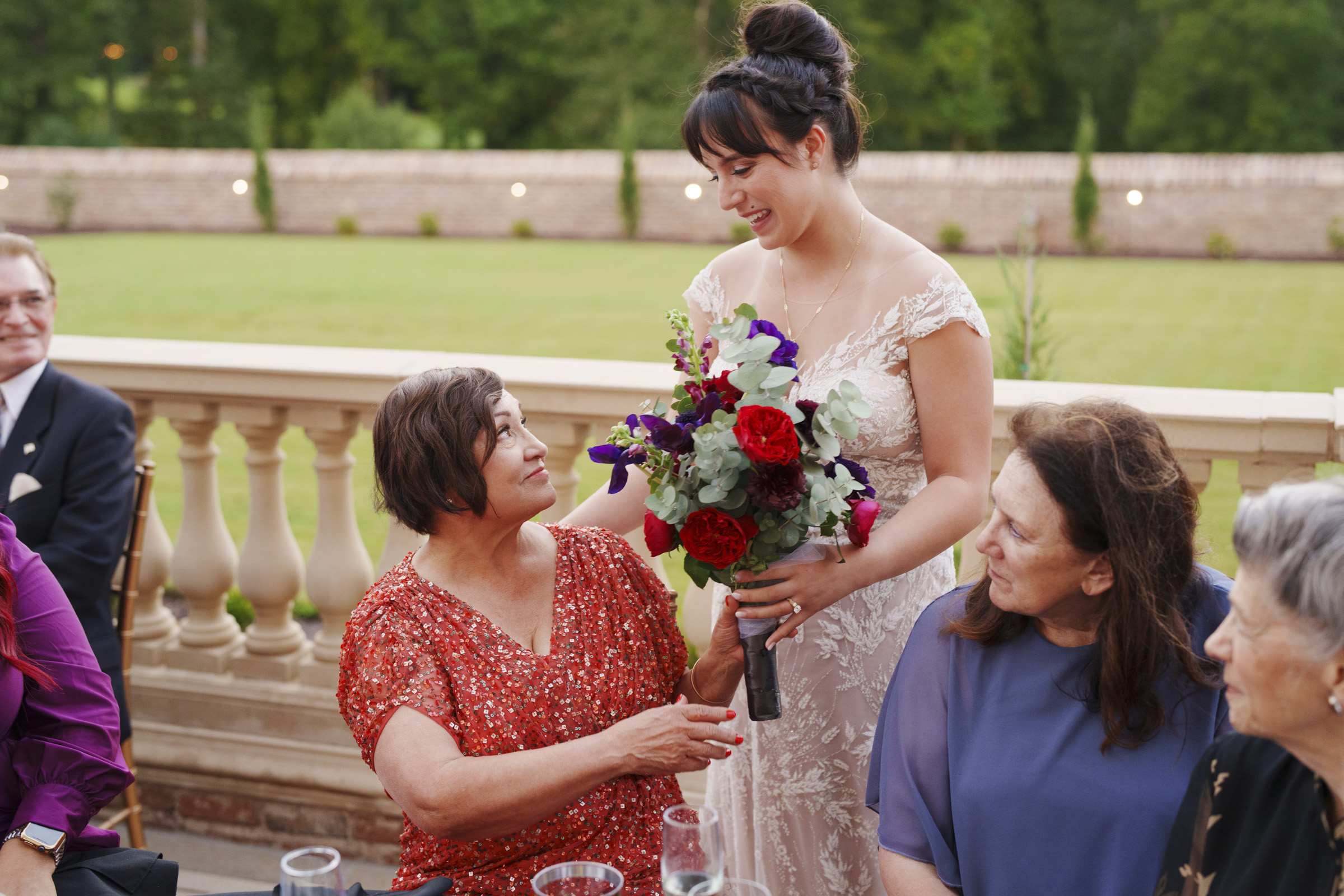 A bride in a lace gown hands a bouquet of red and purple flowers to an older woman in a patterned red dress on the stone terrace of Oxbow Estate. With green lawns and trees as a backdrop, seated guests watch with smiles, captured perfectly by wedding photography.