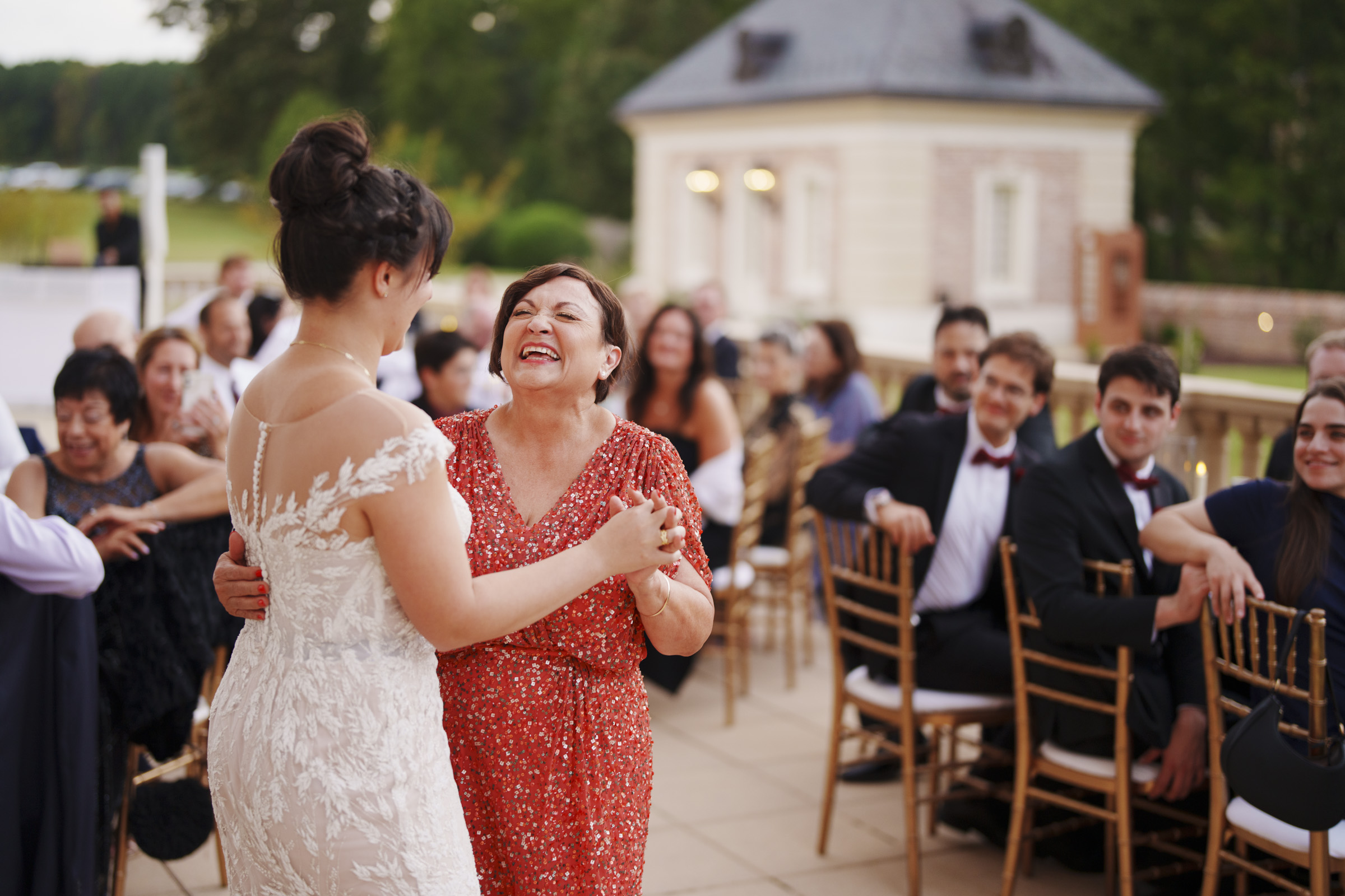 At an Oxbow Estate wedding, a bride in a white dress dances joyfully with a smiling woman in a red dress at an outdoor event. Guests seated on wooden chairs watch and smile, capturing the moment perfectly. A small building and lush green foliage enhance the picturesque setting.