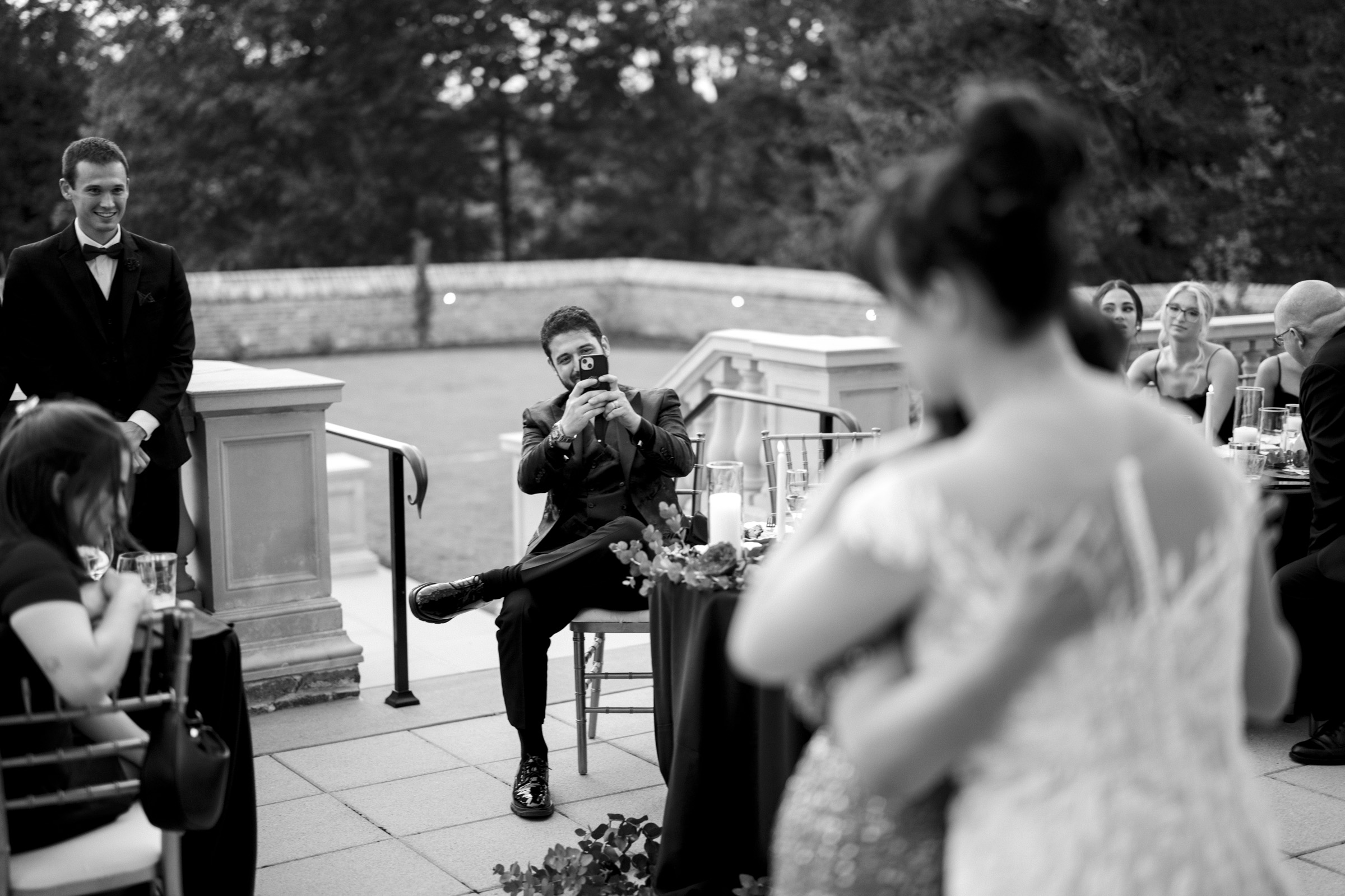 A black-and-white photo captures the elegance of an Oxbow Estate wedding reception. A person is seated, snapping a picture of a couple embracing in the foreground. Guests chat at tables, while a well-dressed man stands smiling on the left. An enchanting outdoor setting completes the scene.