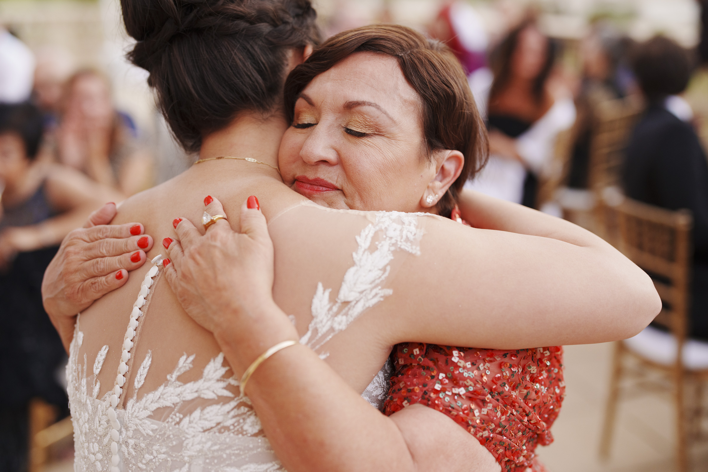 At the Oxbow Estate Wedding, a bride in a white lace gown and a woman in a red sequined dress share an emotional hug. The woman has her eyes closed, displaying a warm and affectionate moment. The background is blurred with seated guests, capturing the essence of heartfelt wedding photography.