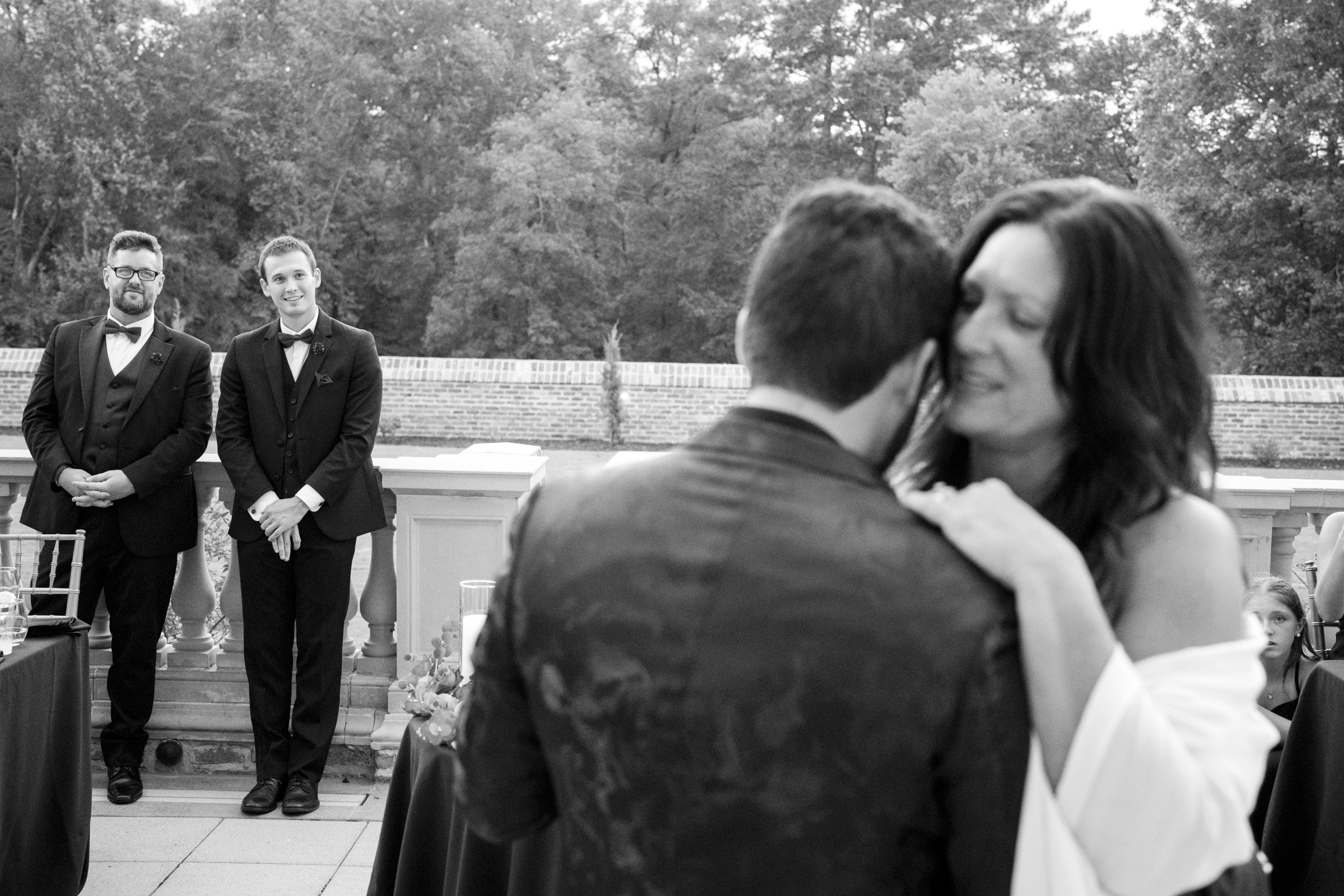 A black-and-white photo captures a couple embracing on a terrace during an Oxbow Estate wedding. Two people in suits stand in the background, observing the scene. Trees and a brick wall can be seen in the distance, adding charm to this intimate moment of wedding photography.