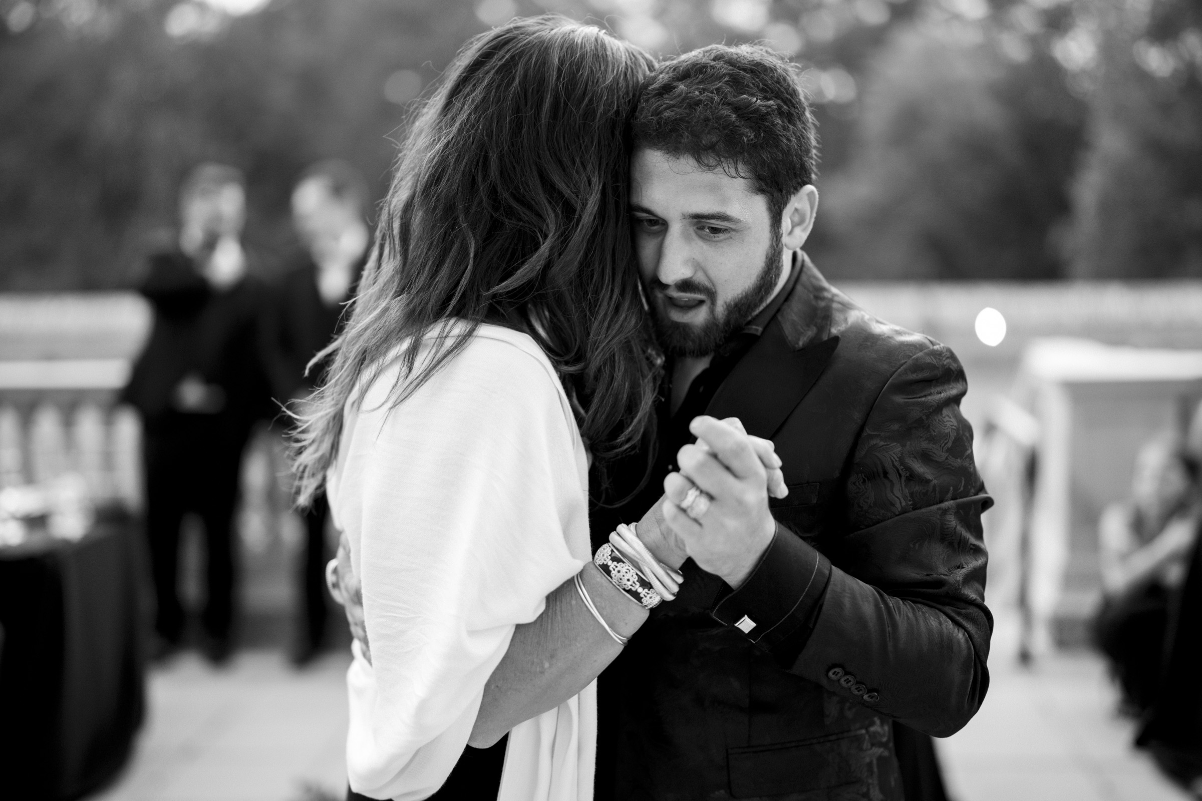 At an Oxbow Estate Wedding, a couple dances closely, their eyes locked in an intimate gaze. The man sports a dark suit while the woman is in light-colored attire. In the background, blurred figures and a railing frame this timeless black and white photo.