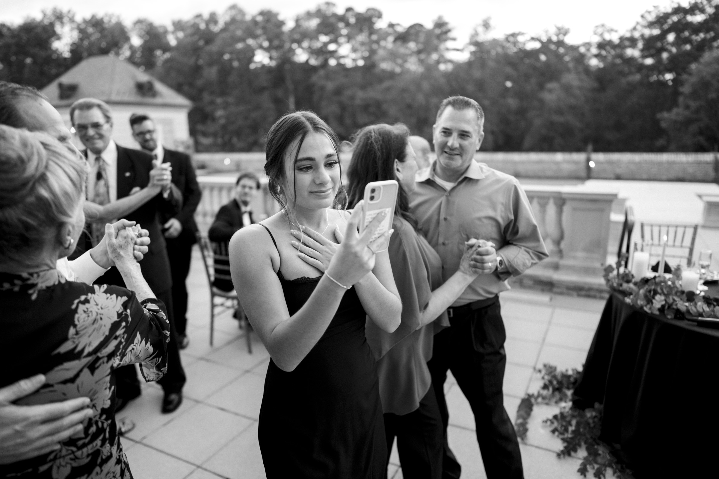 A young woman in a black dress stands on a terrace at Oxbow Estate, holding a phone and appearing emotional. Several people in the background are dancing and smiling, captured beautifully by wedding photography. The outdoor setting is graced with trees lining the horizon.