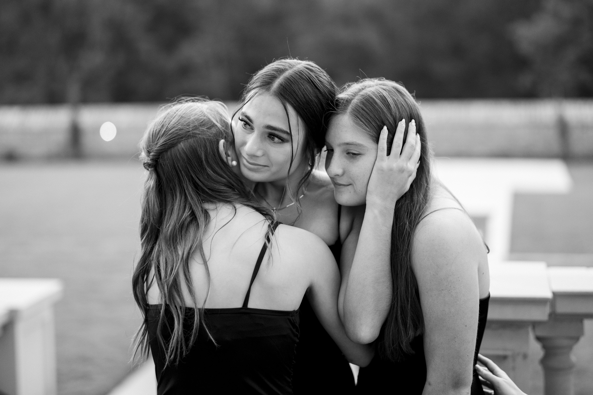 Three young women stand closely together, hugging and comforting each other. Dressed in black against a blurred natural background, the scene exudes tenderness and emotion, reminiscent of moments captured by Oxbow Estate Wedding Photography.