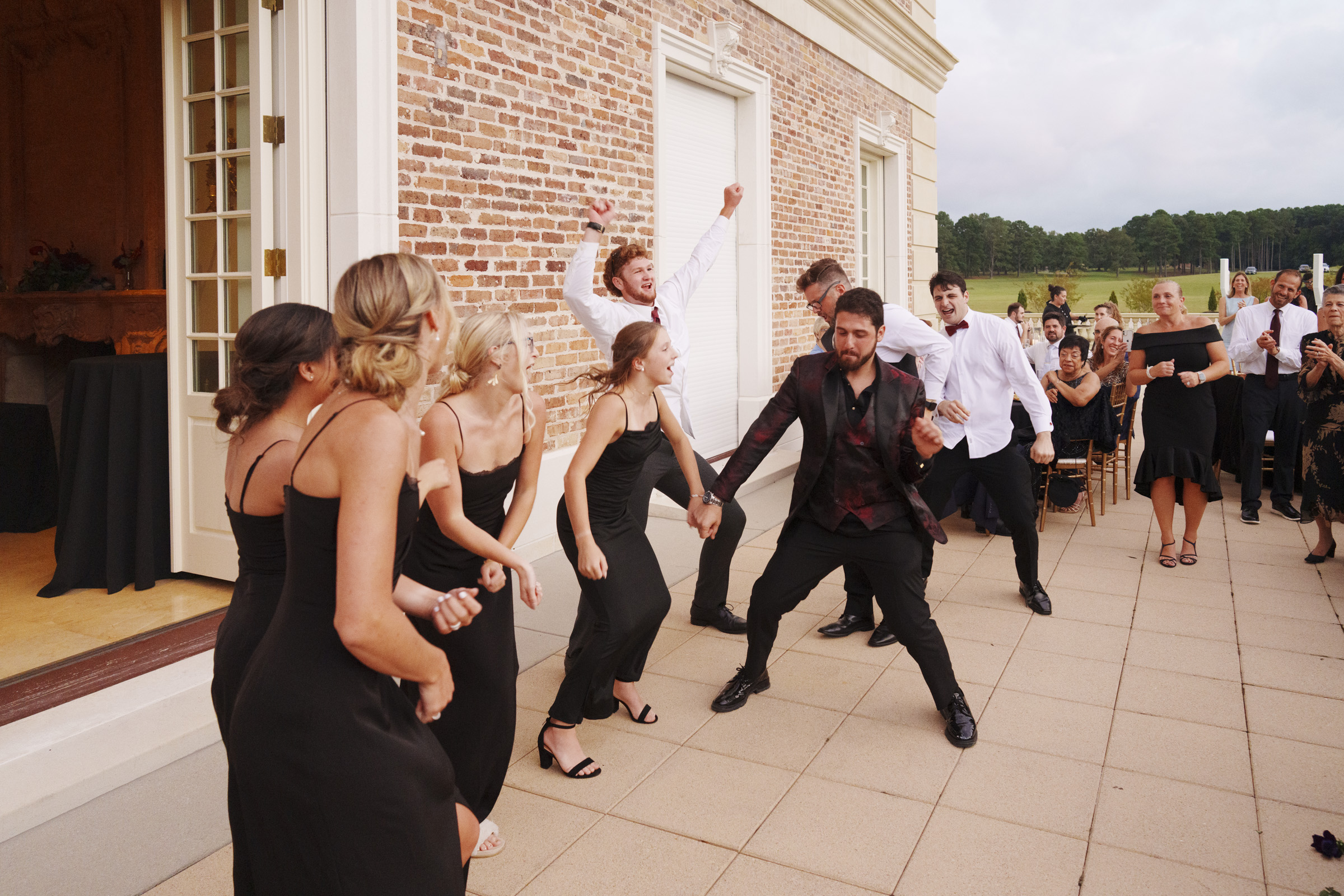 A lively group of people dressed in formal black attire dance energetically on a terrace beside a brick building at an Oxbow Estate wedding. Others in the background watch and enjoy the moment, creating a festive atmosphere that's beautifully captured through photography.