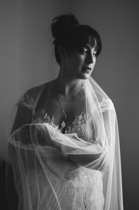 A black and white photo captures a bride at Oxbow Estate, wearing a strapless wedding dress adorned with delicate floral lace. A translucent veil drapes over her as she gazes thoughtfully to the side, her hair elegantly styled in an updo.