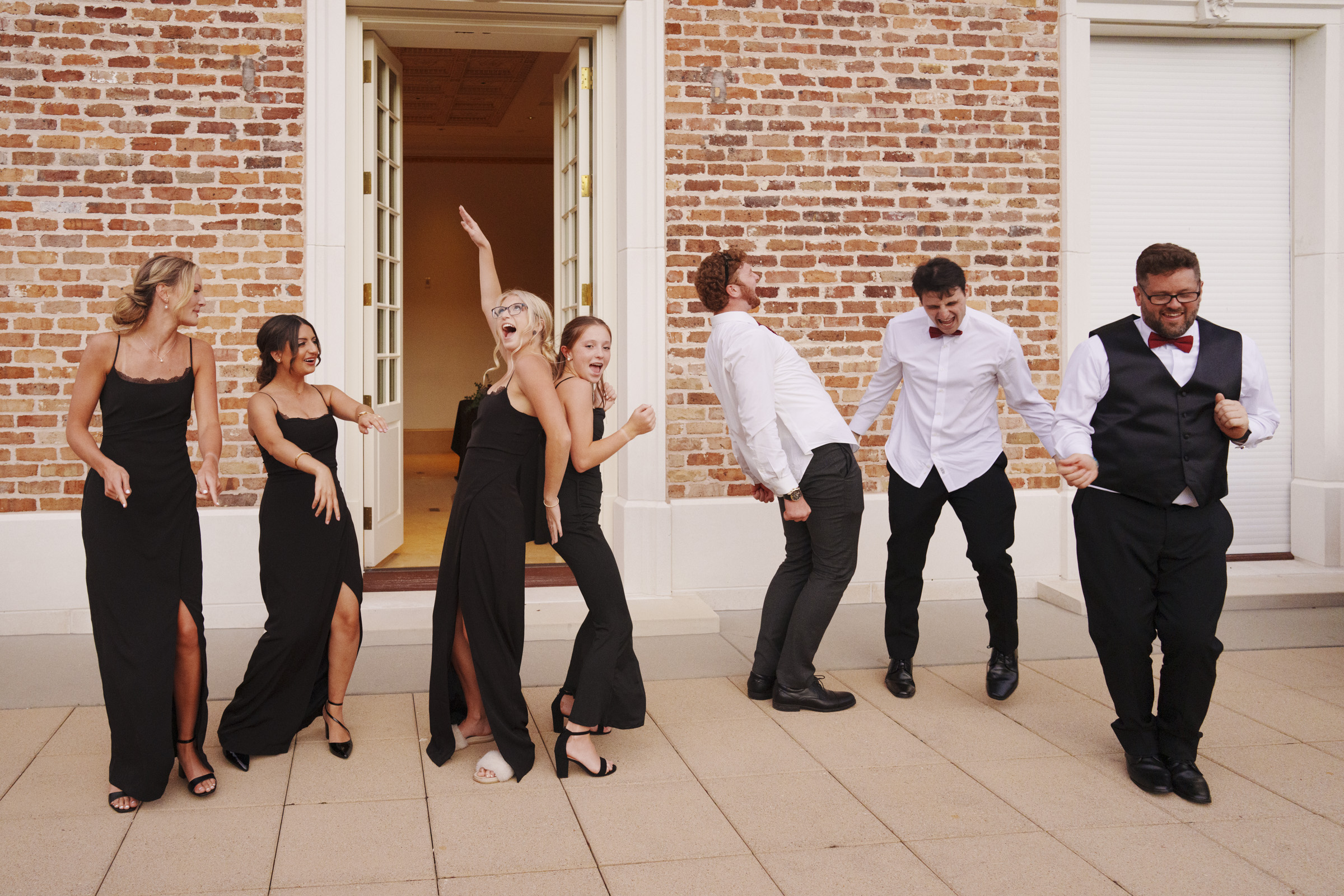 A group of people dressed in formal attire are joyfully posing and dancing in front of a brick building at Oxbow Estate. The women wear black dresses, while the men don white shirts or black suits with bow ties—capturing a moment of wedding photography bliss.