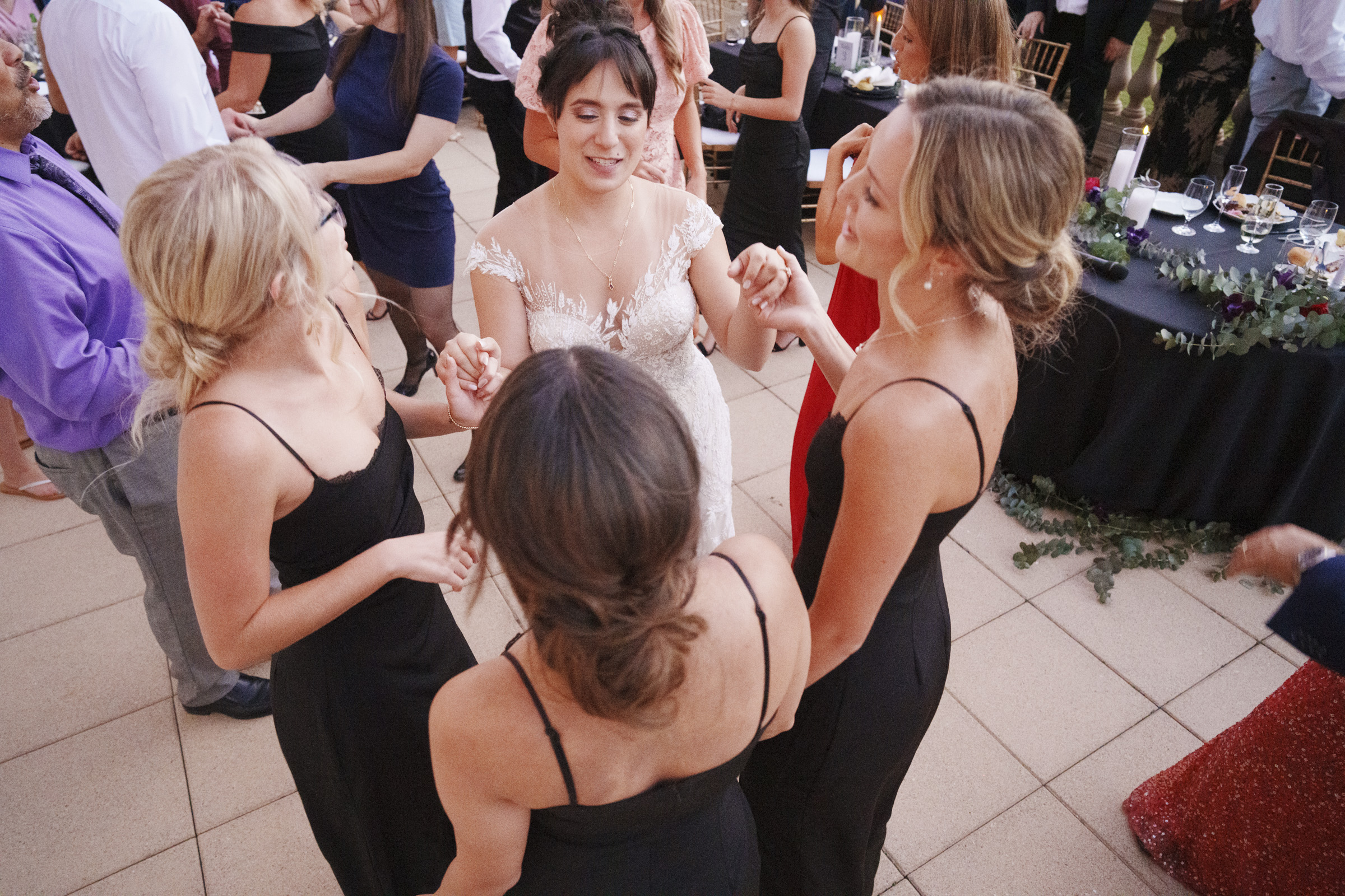 At the Oxbow Estate, a bride in a white gown joyfully dances with her four bridesmaids in black dresses at the wedding reception. They hold hands in a circle, surrounded by other guests on the elegant patio, as captured beautifully by the wedding photography team.