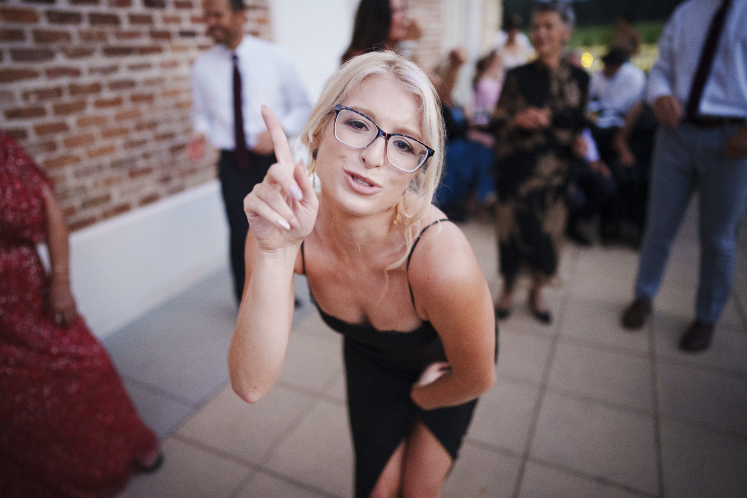 A woman with blonde hair and glasses, in a black dress, gestures playfully surrounded by smiling guests at an Oxbow Estate wedding. Snapshots of joy unfold against the charming brick wall backdrop.