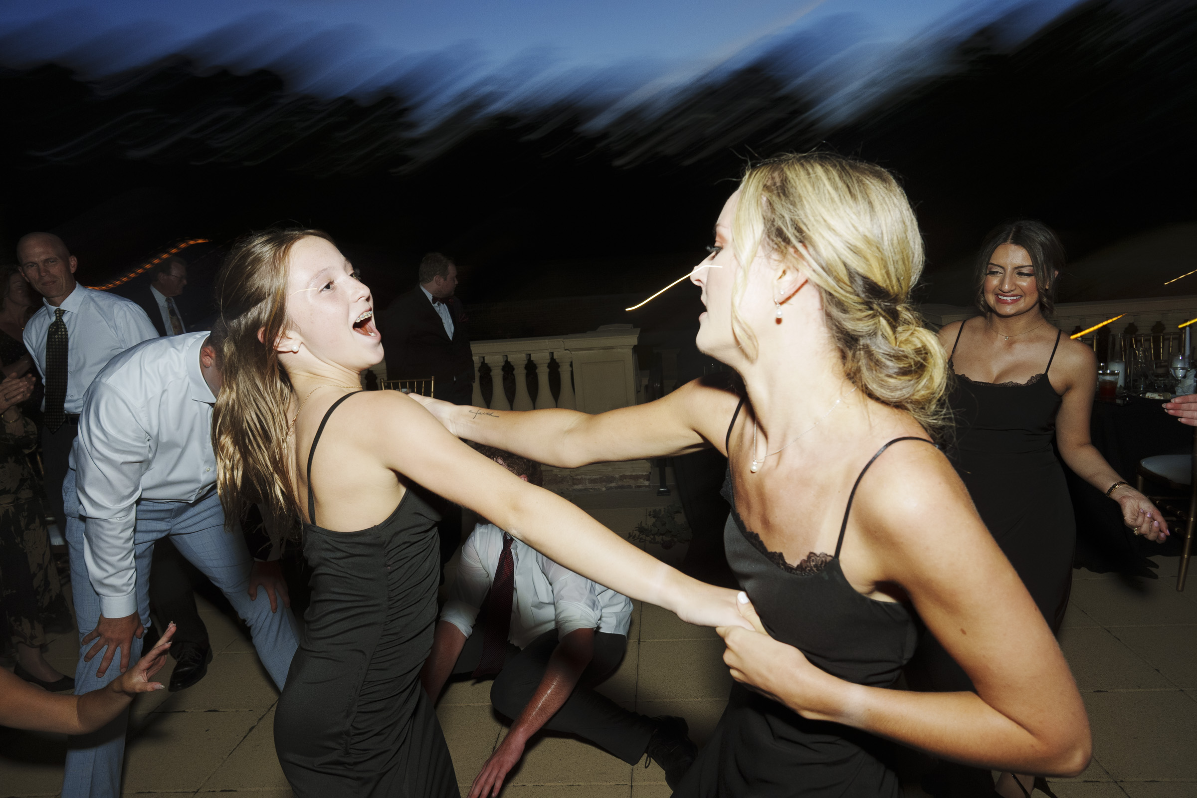 At the Oxbow Estate, the night came alive with vibrant energy as two women in black dresses joyfully danced under the stars. One spun gracefully while others in formal attire swayed to the rhythm, capturing a vivid moment of wedding photography magic.