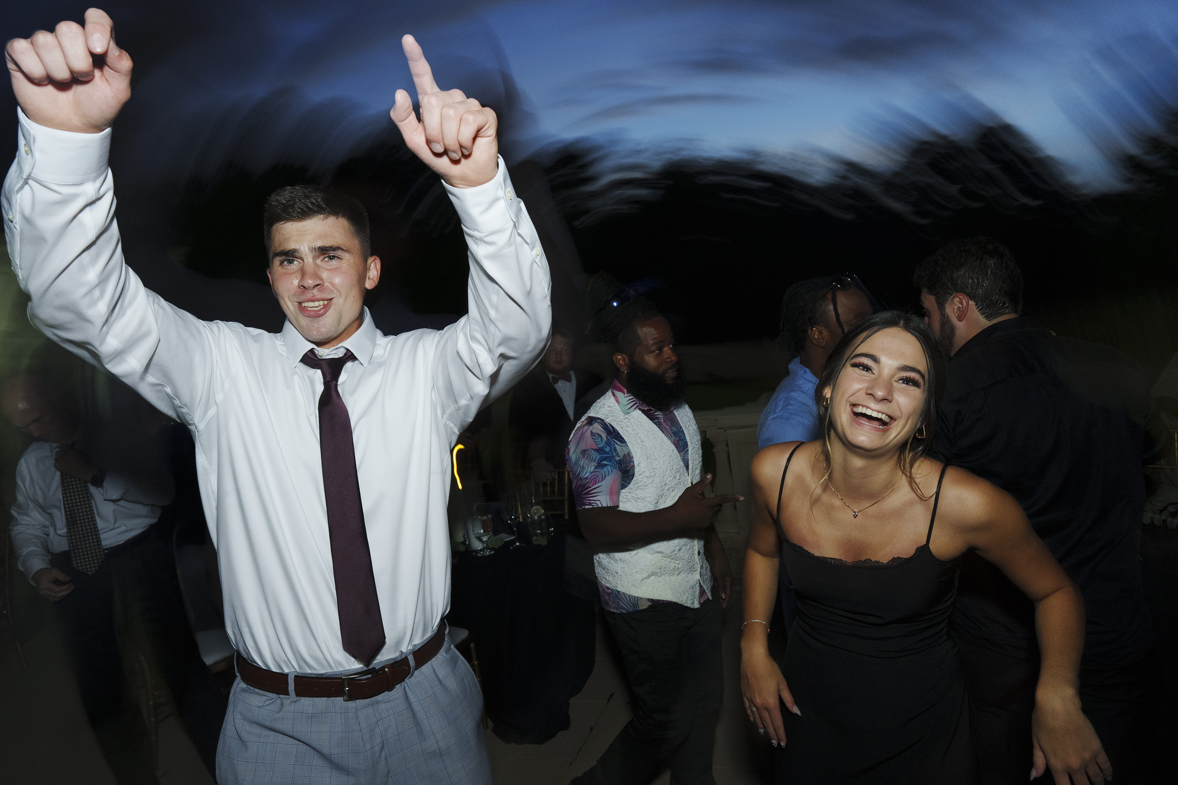 At an Oxbow Estate wedding, a man and woman dance energetically at the party. The man in a white shirt and tie has his hands raised, while the woman in a black dress smiles broadly. The background shows blurred figures and trees, capturing the lively essence of the outdoor celebration.