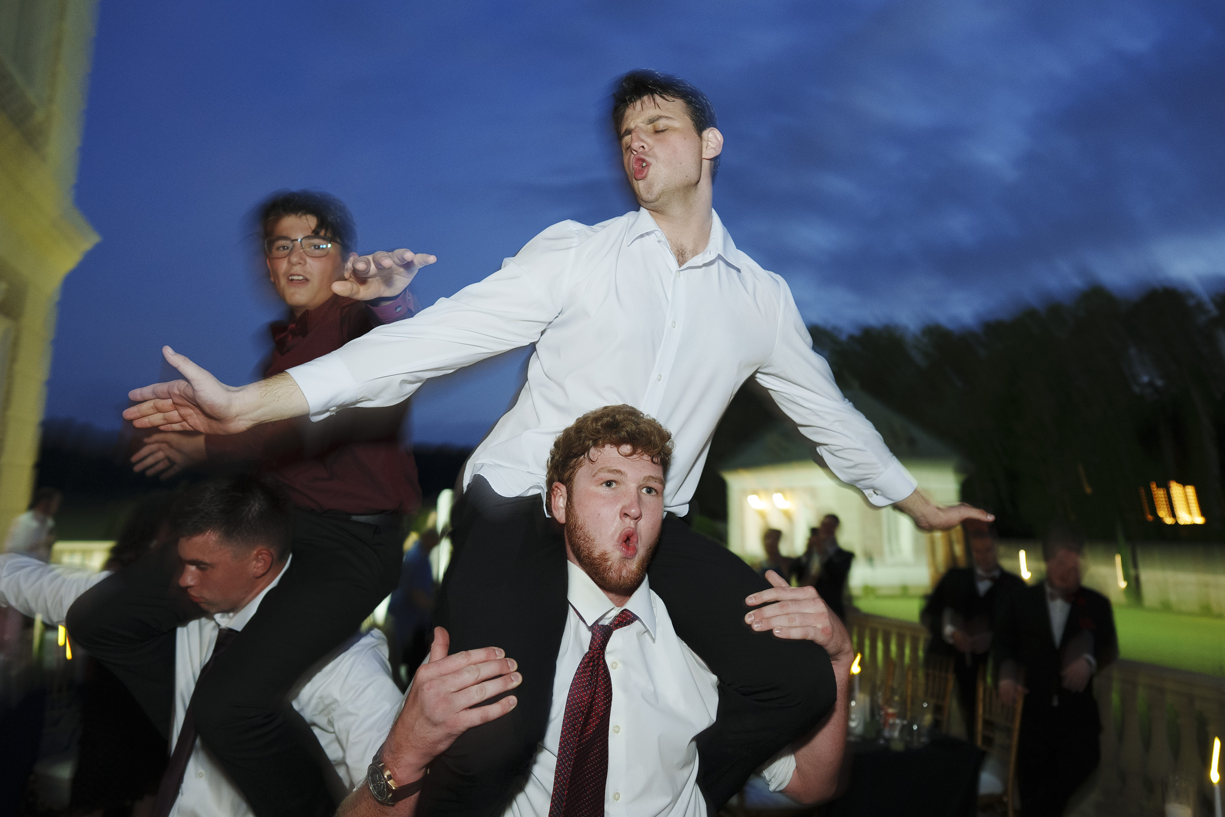 At an Oxbow Estate wedding, four young men, two perched on each other's shoulders, strike playful poses at the evening event. Clad in formal attire with ties, they stand against a darkening sky and blurred greenery, capturing the joyous spirit of the occasion through photography.