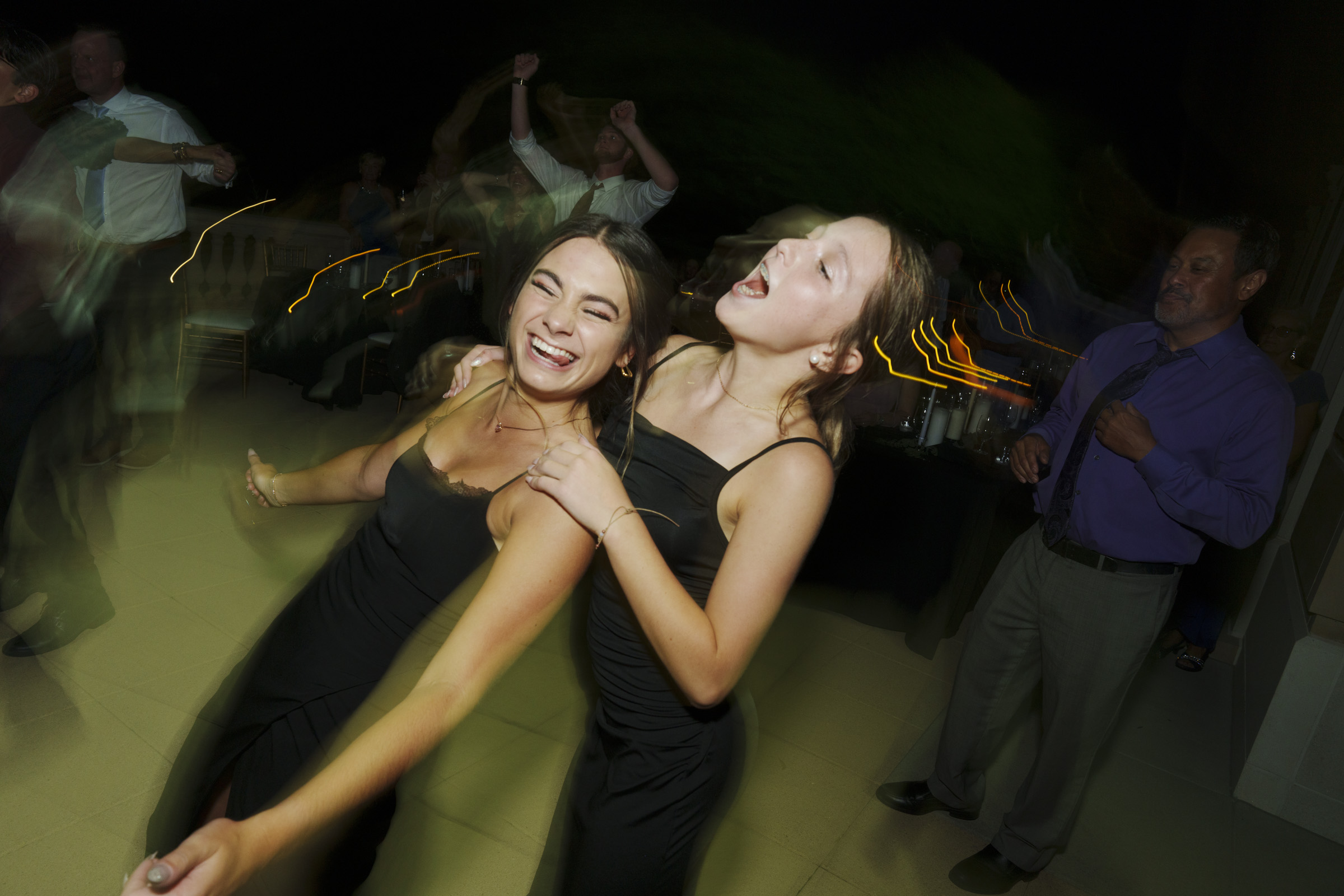 Two people in black dresses joyfully dance together with their arms around each other at a lively party. The dimly lit background, reminiscent of Oxbow Estate Wedding Photography, frames other guests, enhancing the festive atmosphere.