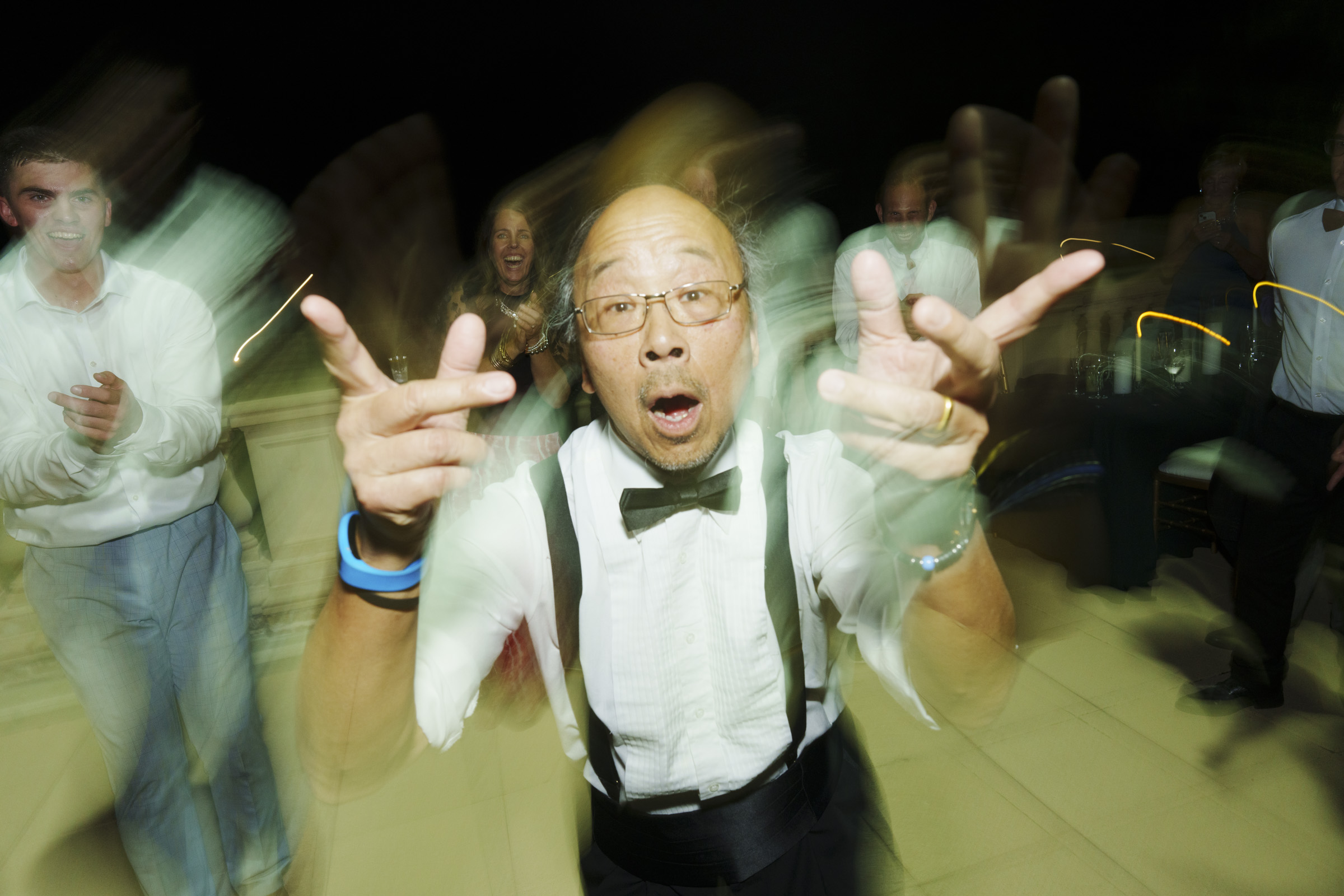 A man wearing a bow tie and suspenders dances energetically at the Oxbow Estate wedding, with motion blur accentuating his lively moves. In the background, guests in formal attire join the celebration, set against a dark backdrop that highlights the joyful ambiance of the event.