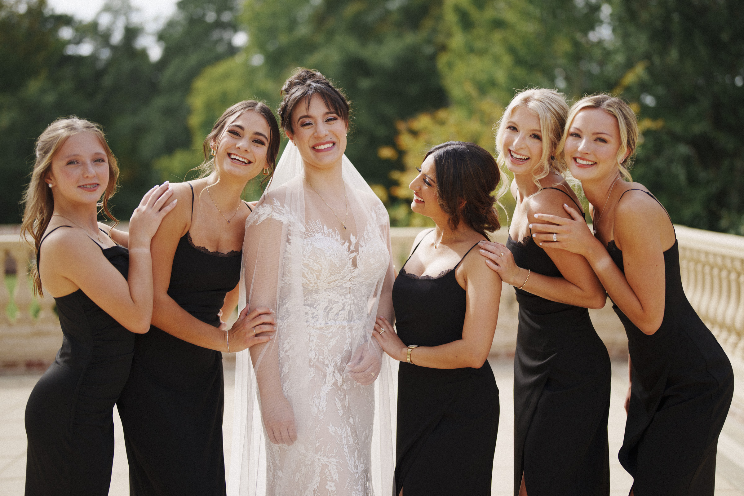 At the picturesque Oxbow Estate, a bride in a white gown and veil beams with her five bridesmaids in matching black dresses, posing elegantly on an outdoor terrace with a backdrop of lush trees.