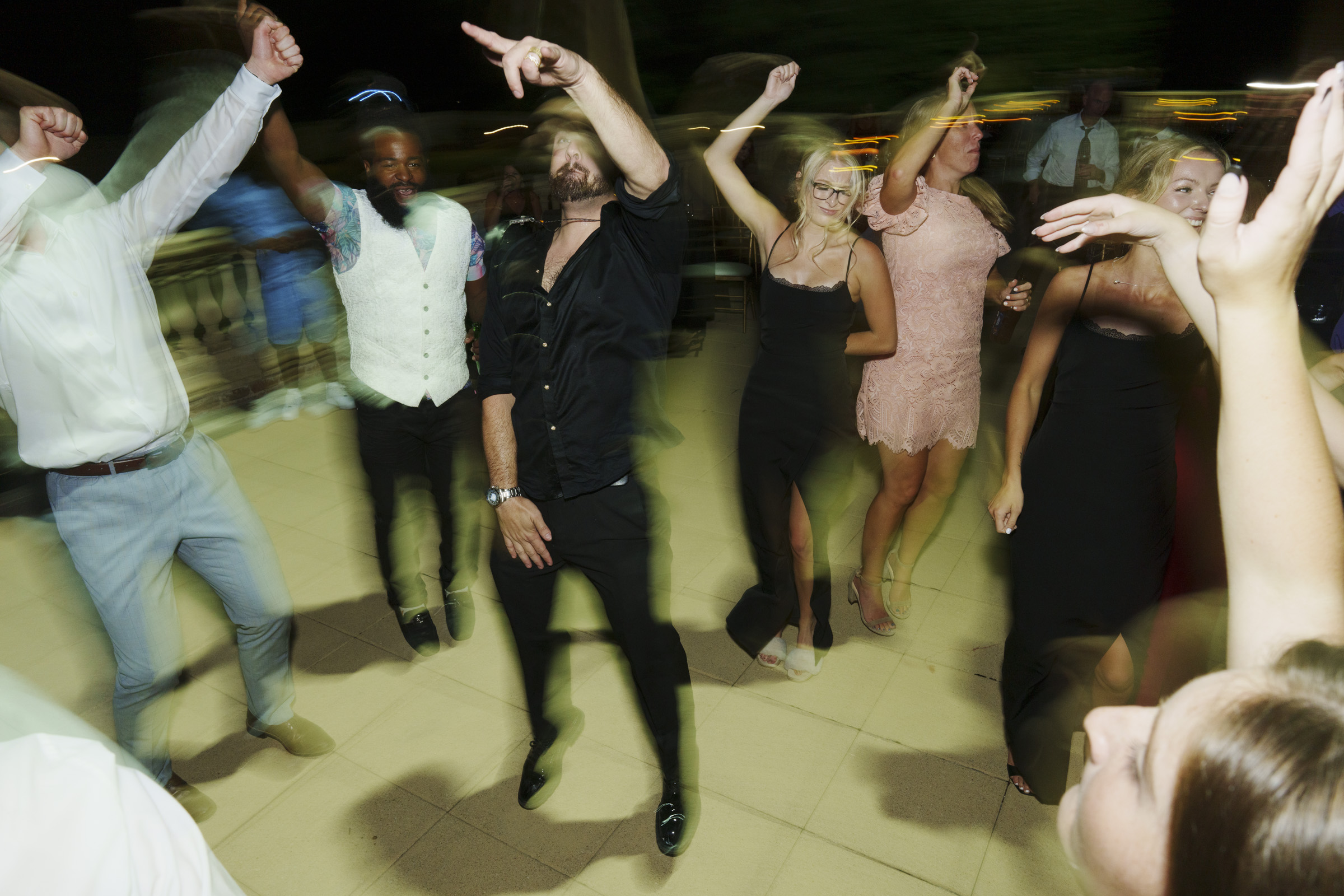 A group of people dancing energetically at a party on an outdoor patio at night, with blurred motion capturing the lively atmosphere. Most are dressed in formal or semi-formal attire at the Oxbow Estate wedding celebration, enjoying every moment.