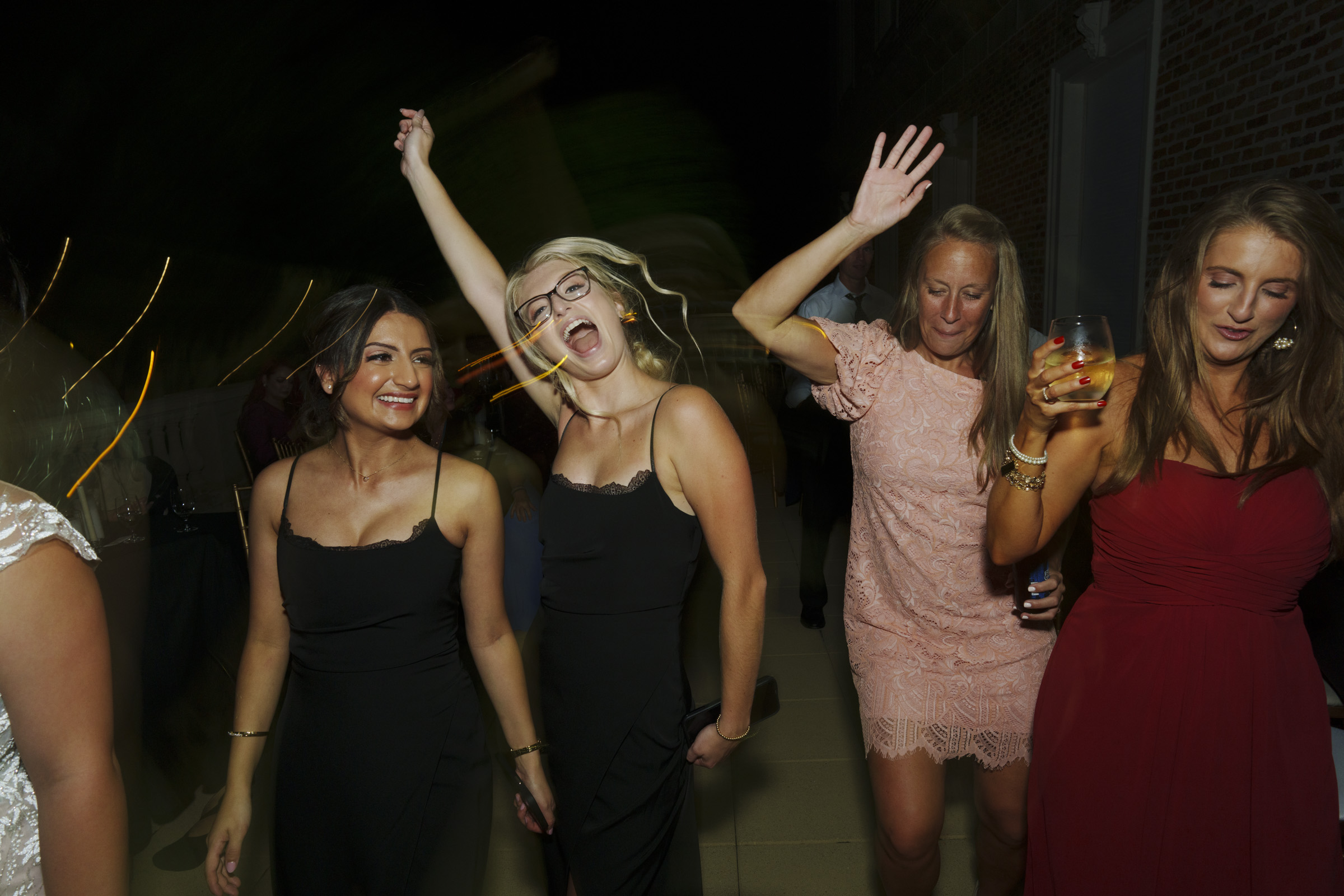 A group of women is dancing joyfully at a nighttime event, captured beautifully in Oxbow Estate Wedding Photography. They are wearing elegant dresses, two with hands raised, one holding a drink. The background is dimly lit, enhancing the lively atmosphere.