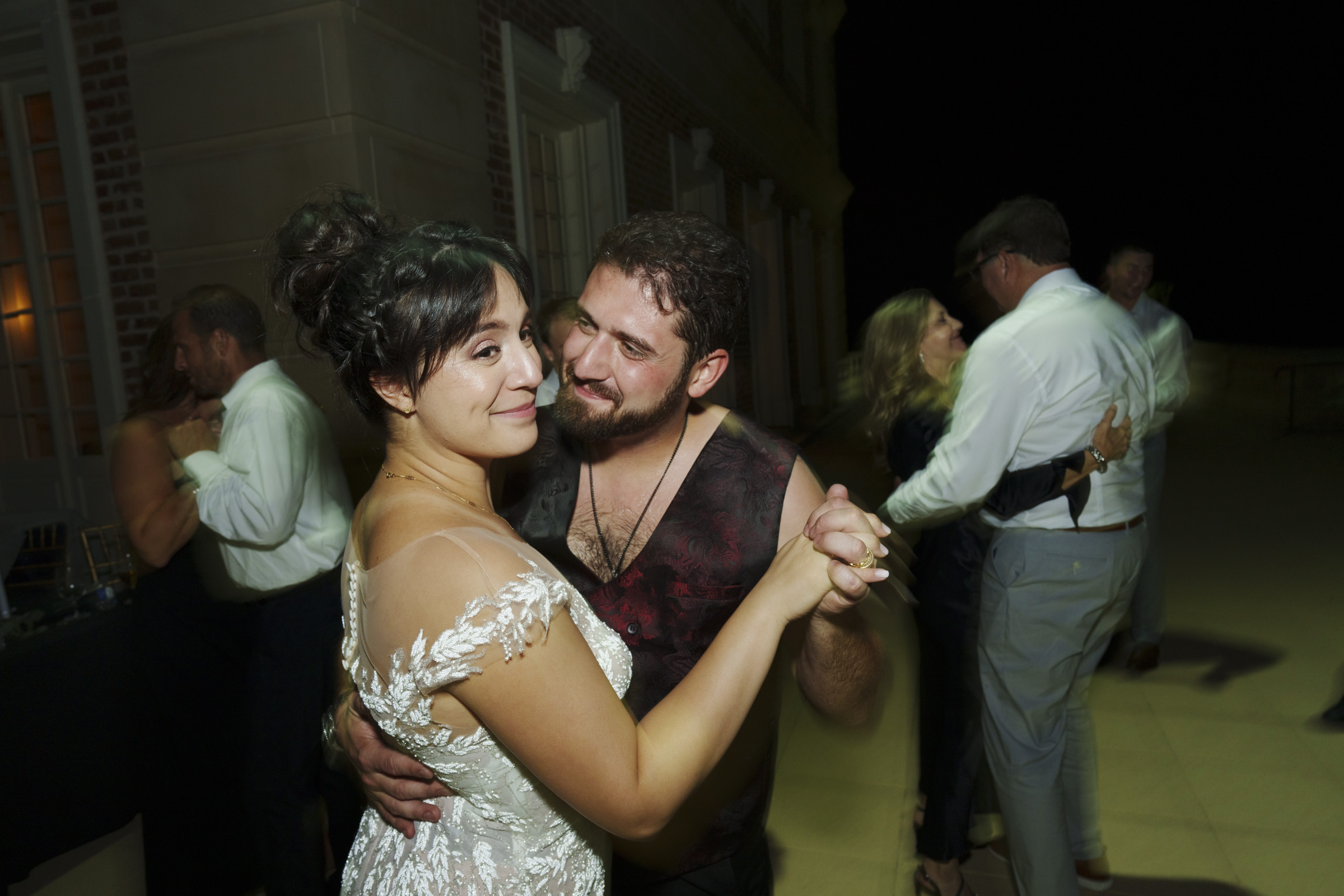 At an enchanting Oxbow Estate wedding, a couple sways closely under the starlit sky. The woman, in a flowing white dress, and the man, donning a dark, patterned vest, create a picture-perfect moment. Other couples join in the dance as the picturesque backdrop adds to the evening's magic.