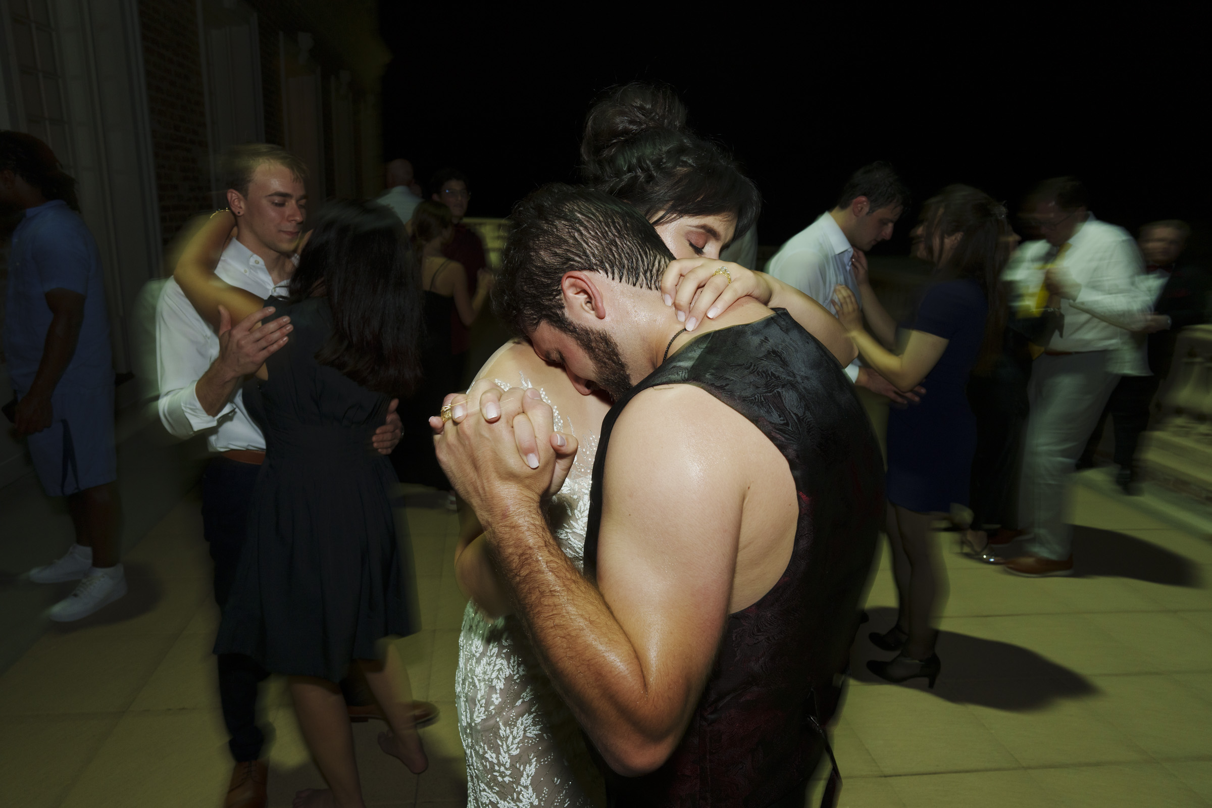 A couple dances closely on an outdoor terrace at night, surrounded by other dancing pairs. Under the gaze of Oxbow Estate Wedding Photography, the man in a dark vest and the woman in a white dress are set against warmly dressed guests in a softly lit setting.