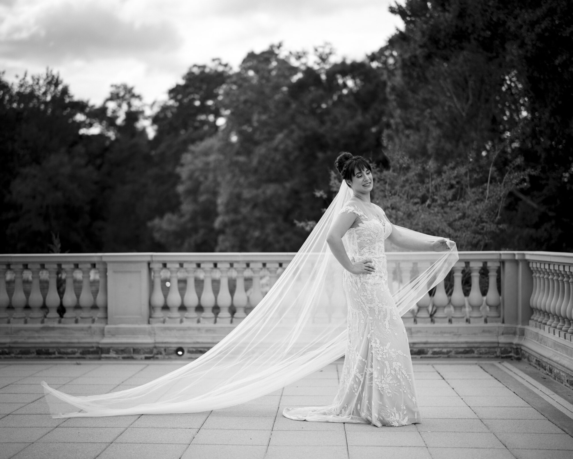 A bride in a lace gown and long veil stands on the terrace of Oxbow Estate, posing elegantly against a backdrop of trees. The black and white image highlights the intricate details of her dress and flowing veil, capturing the essence of estate wedding photography at its finest.