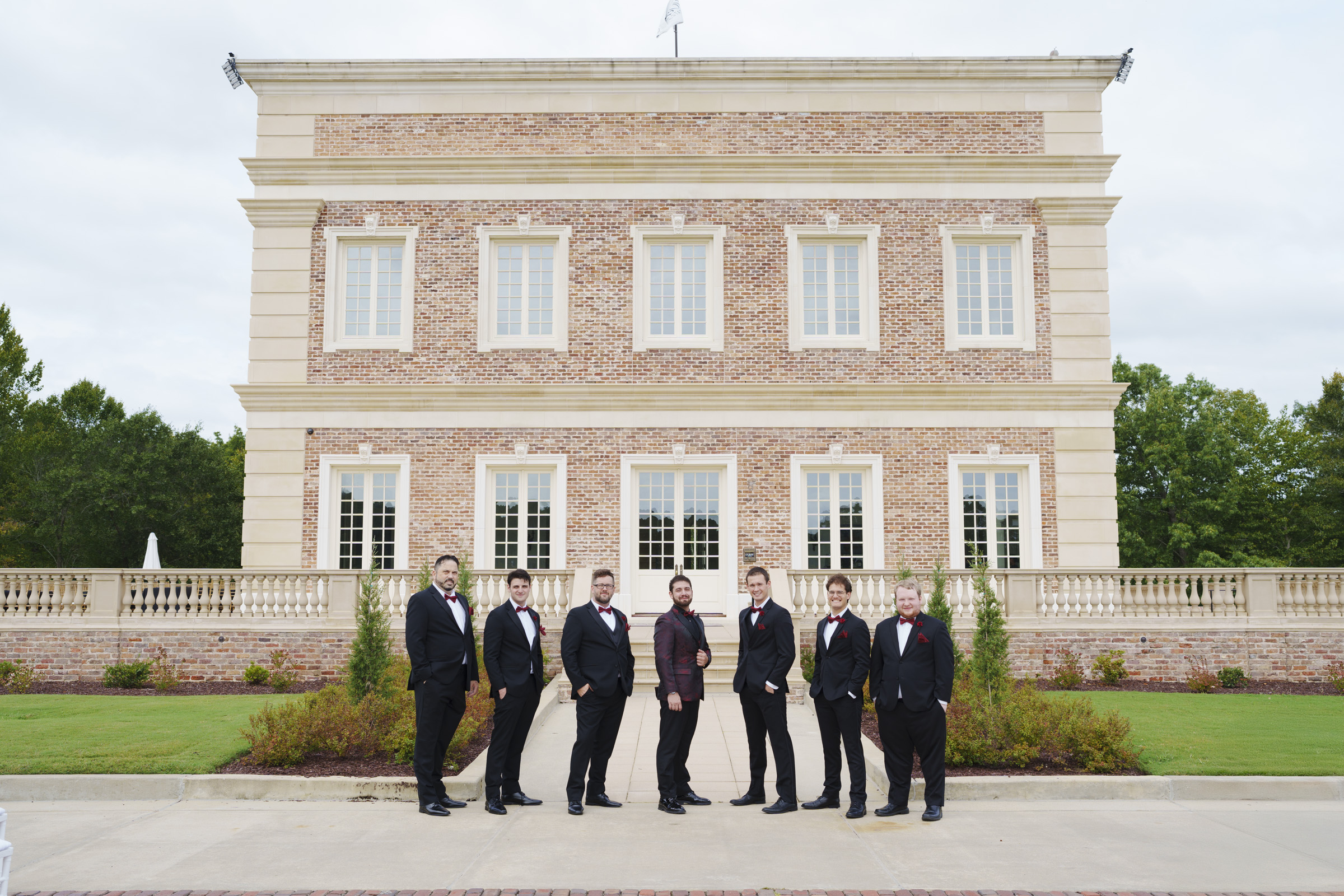 Seven men in formal suits stand in a line outside the grand, classical-style brick building of Oxbow Estate. The overcast sky adds a dramatic touch, while the lush greenery completes this perfect scene for wedding photography.