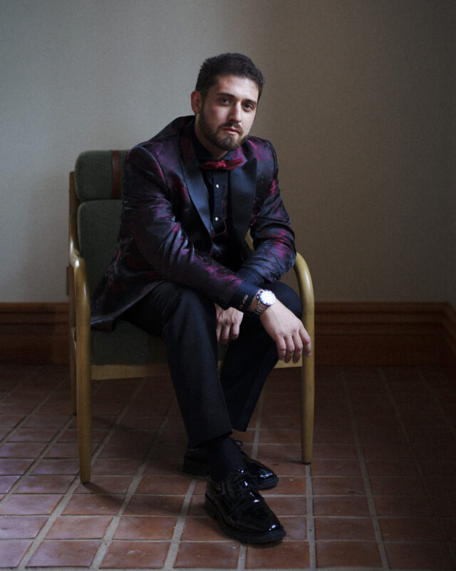 A man sits in a chair, wearing a dark, patterned suit with a red bow tie and shiny black shoes. He has a watch on his left wrist, posed gracefully for an Oxbow Estate Wedding Photography session in a well-lit brick-floored room.