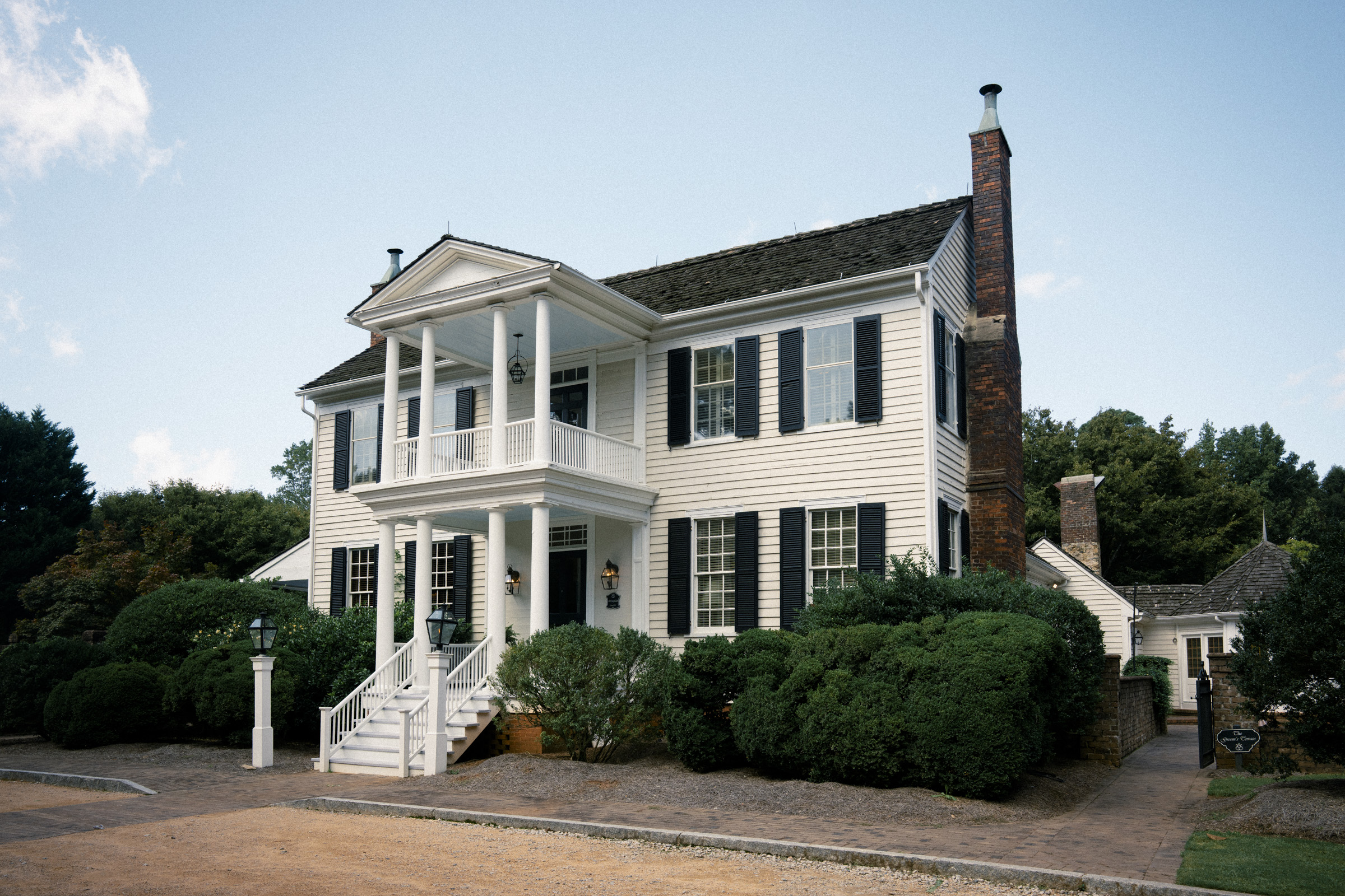 A white two-story colonial-style house with black shutters, a brick chimney, and a front porch supported by columns. It is surrounded by green bushes and set against a blue sky. A paved walkway leads to the entrance.