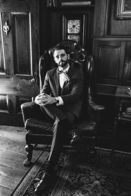 A man in a suit and bow tie sits in a large leather armchair, hands clasped. The room is wood-paneled with bookshelves, an old clock, and a closed door. The setting is elegant and vintage. The image is in black and white.