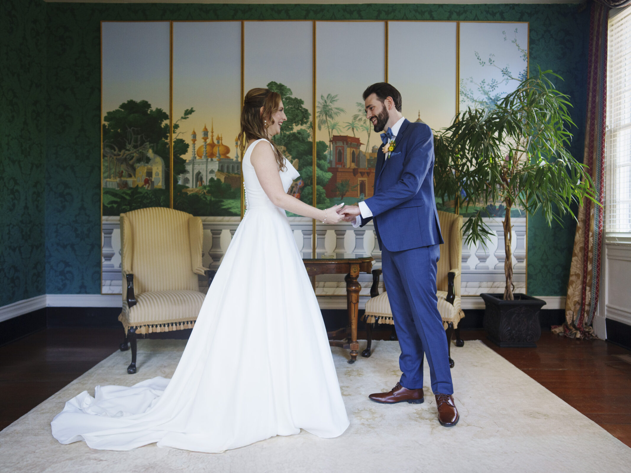 A bride in a white dress and a groom in a blue suit hold hands and smile at each other. They stand in a decorated room with a painted backdrop and green walls, flanked by two chairs and a large plant.