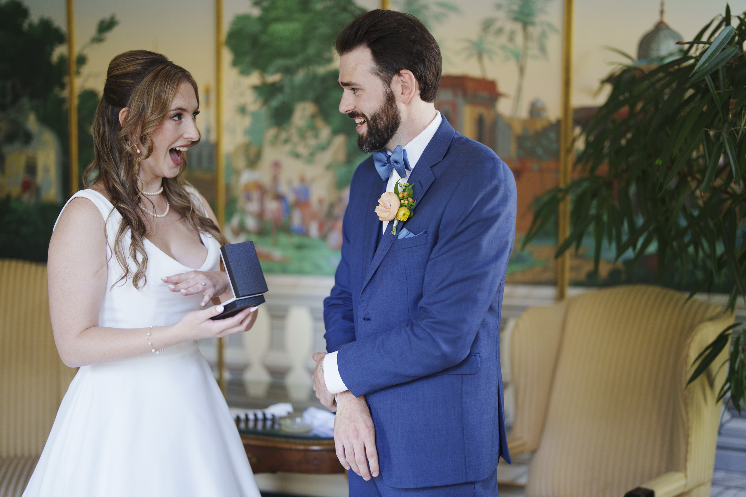 A bride in a white dress, surprised and joyful, holds a jewelry box. A groom in a blue suit and bow tie watches her with a smile. They stand indoors with elegant decor, including a large plant and a mural in the background.