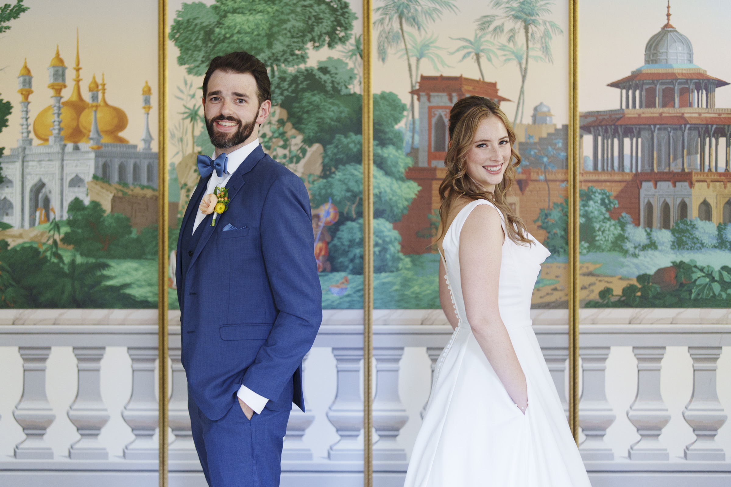 A bride and groom stand back to back, both smiling. The groom is in a blue suit with a yellow boutonniere, and the bride is in a white dress. They are in front of a colorful mural depicting architectural scenes and greenery.